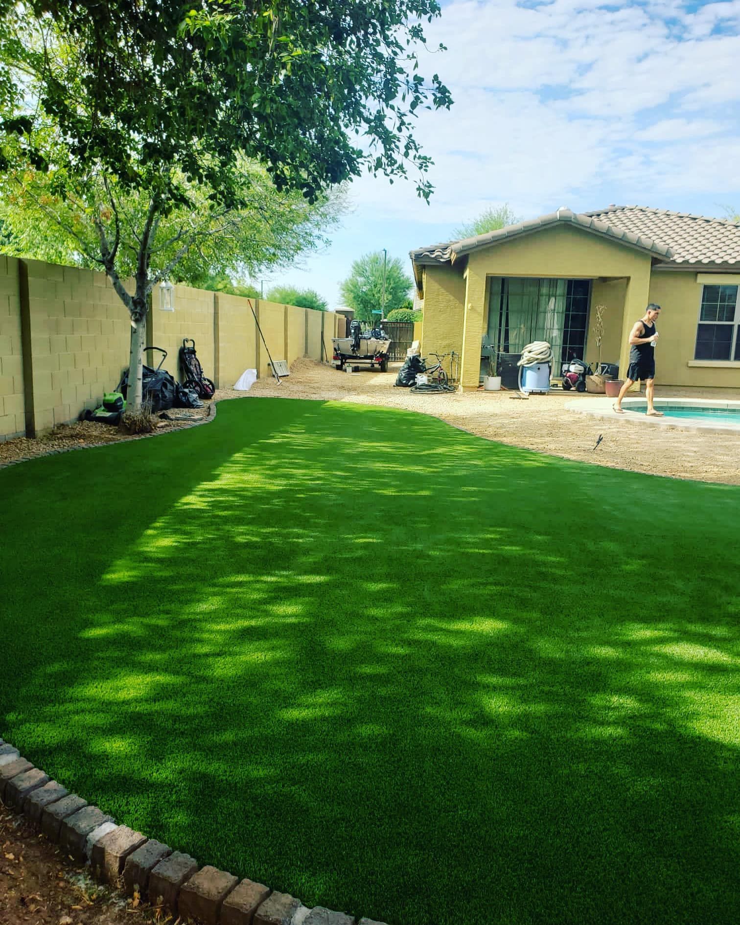 A lush green artificial lawn bordered by pavers, next to a house with a pool and a person walking in the background.