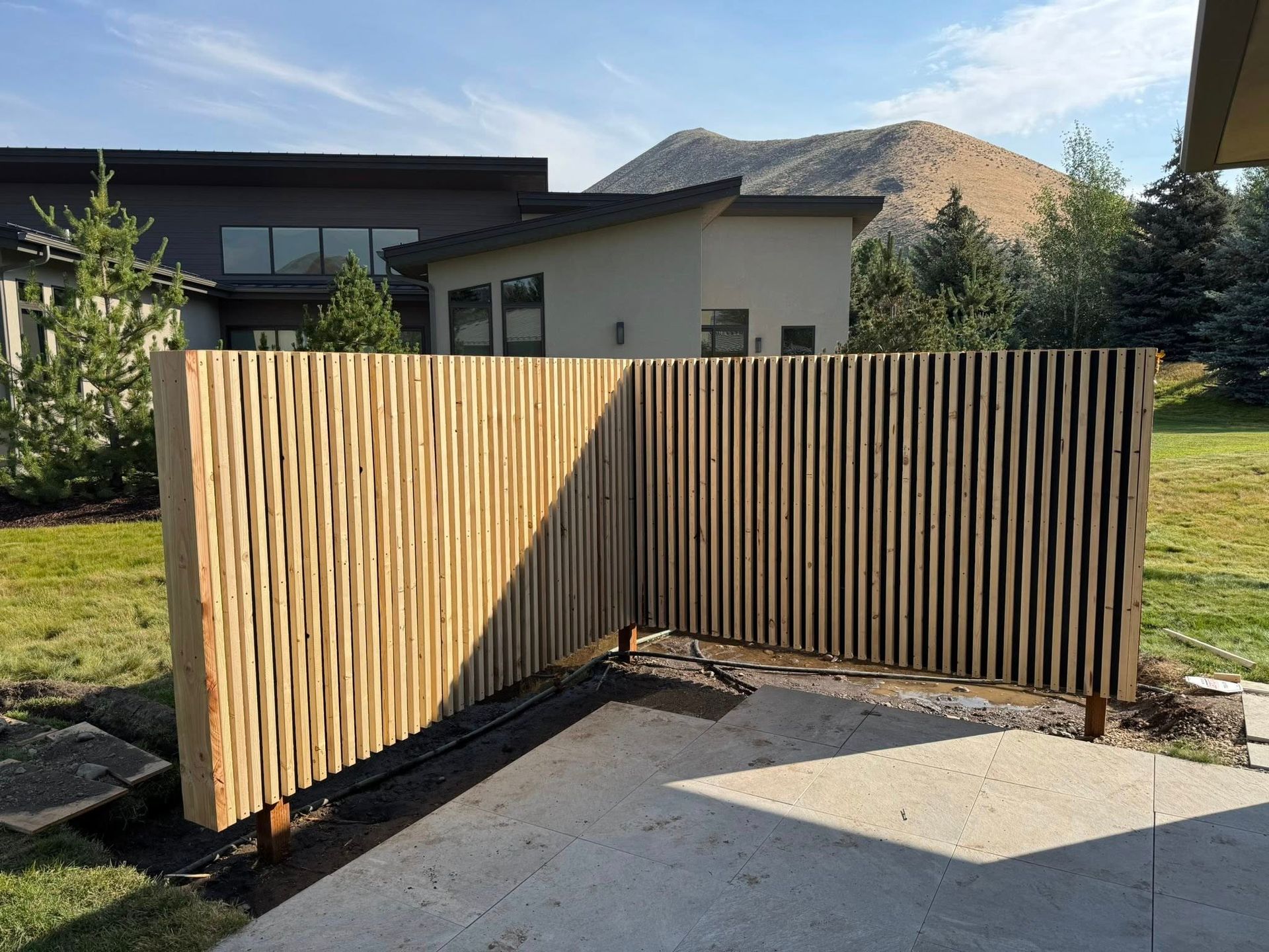 Wooden privacy fence with vertical slats and carved wavy panels on a patio, houses and mountain in the background.
