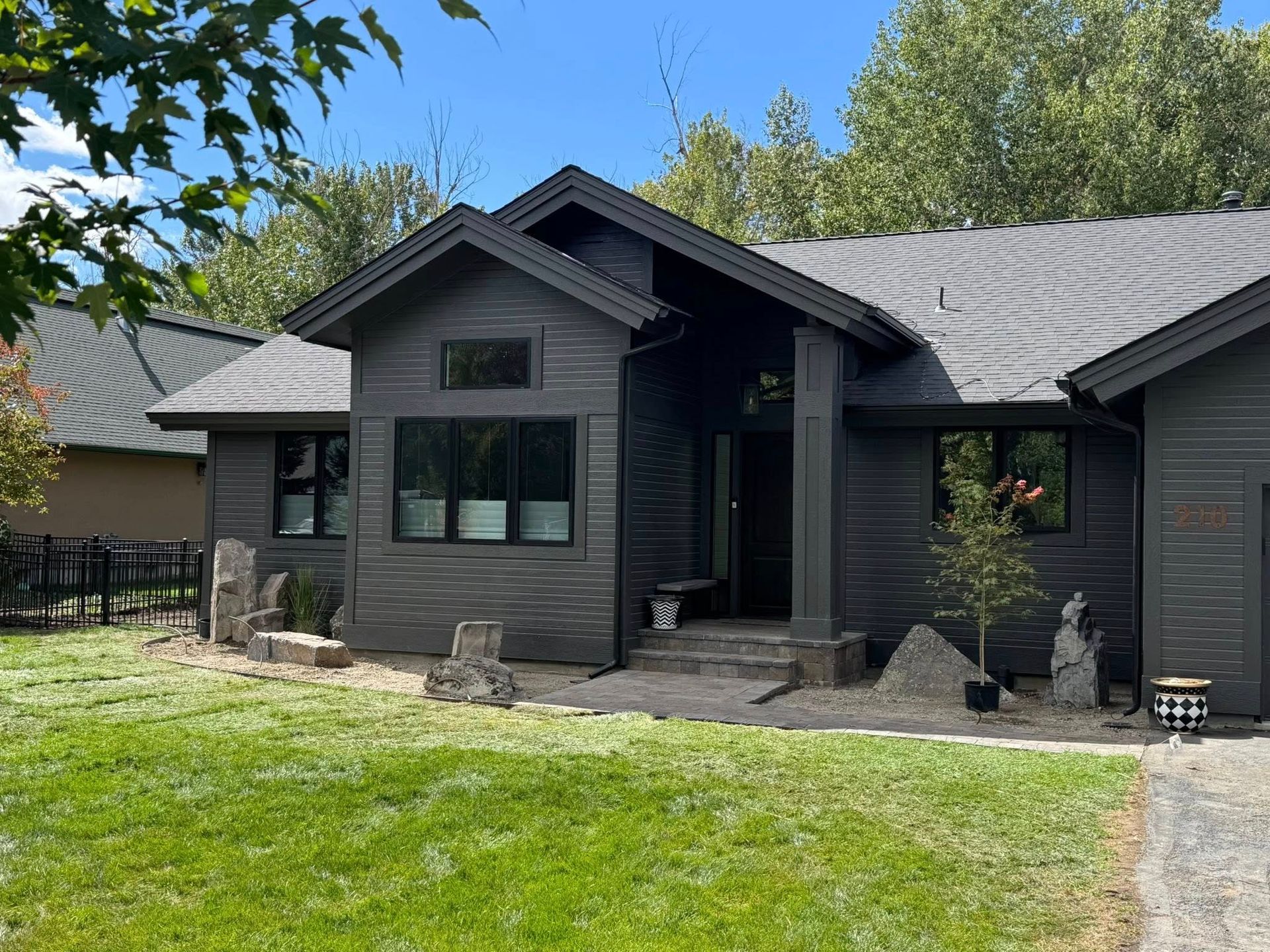 Dark gray house with black-framed windows, surrounded by green grass and trees on a sunny day.