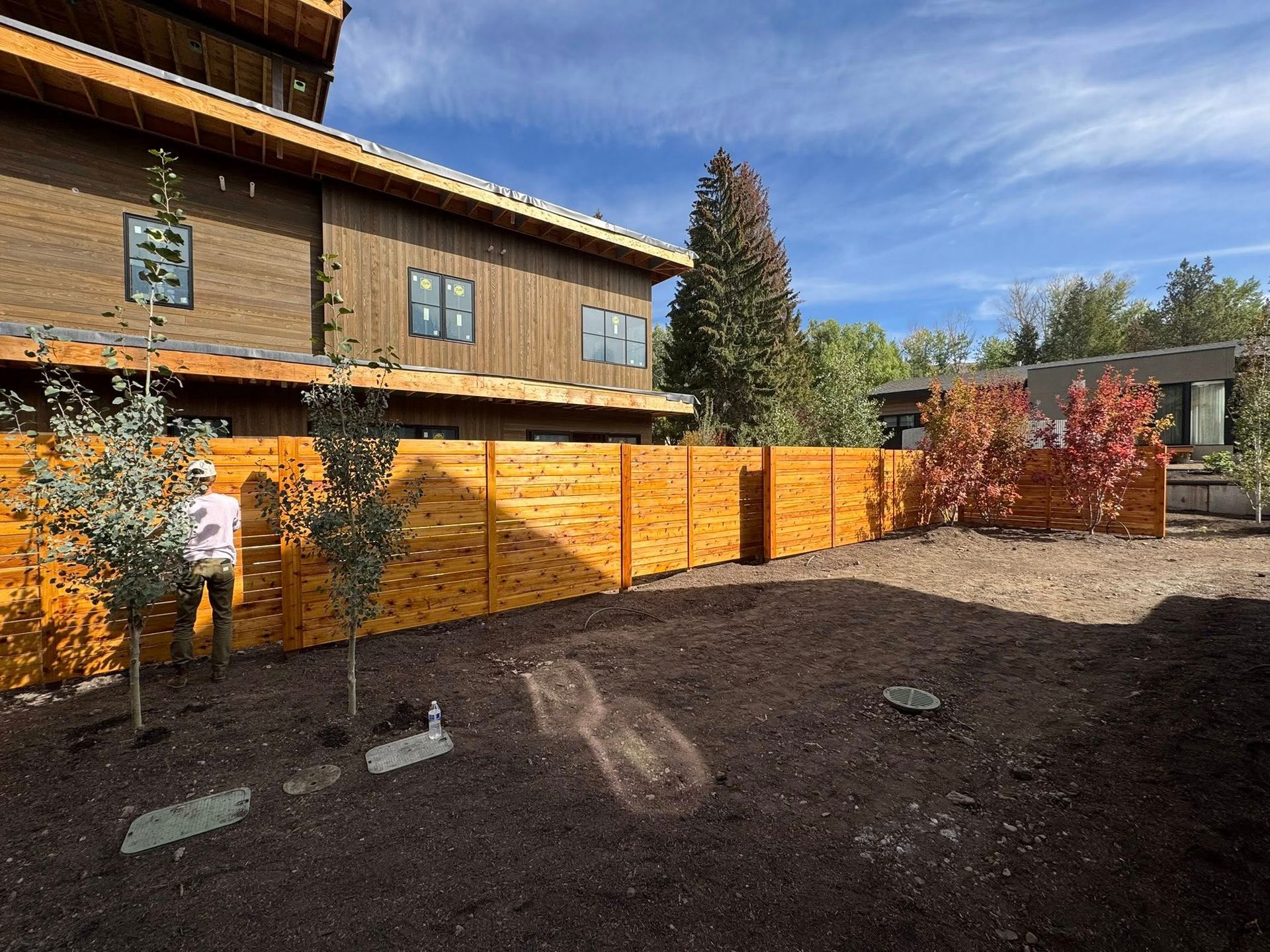 Wooden fence surrounds a yard with a two-story brown house and a smaller building visible. Blue sky.