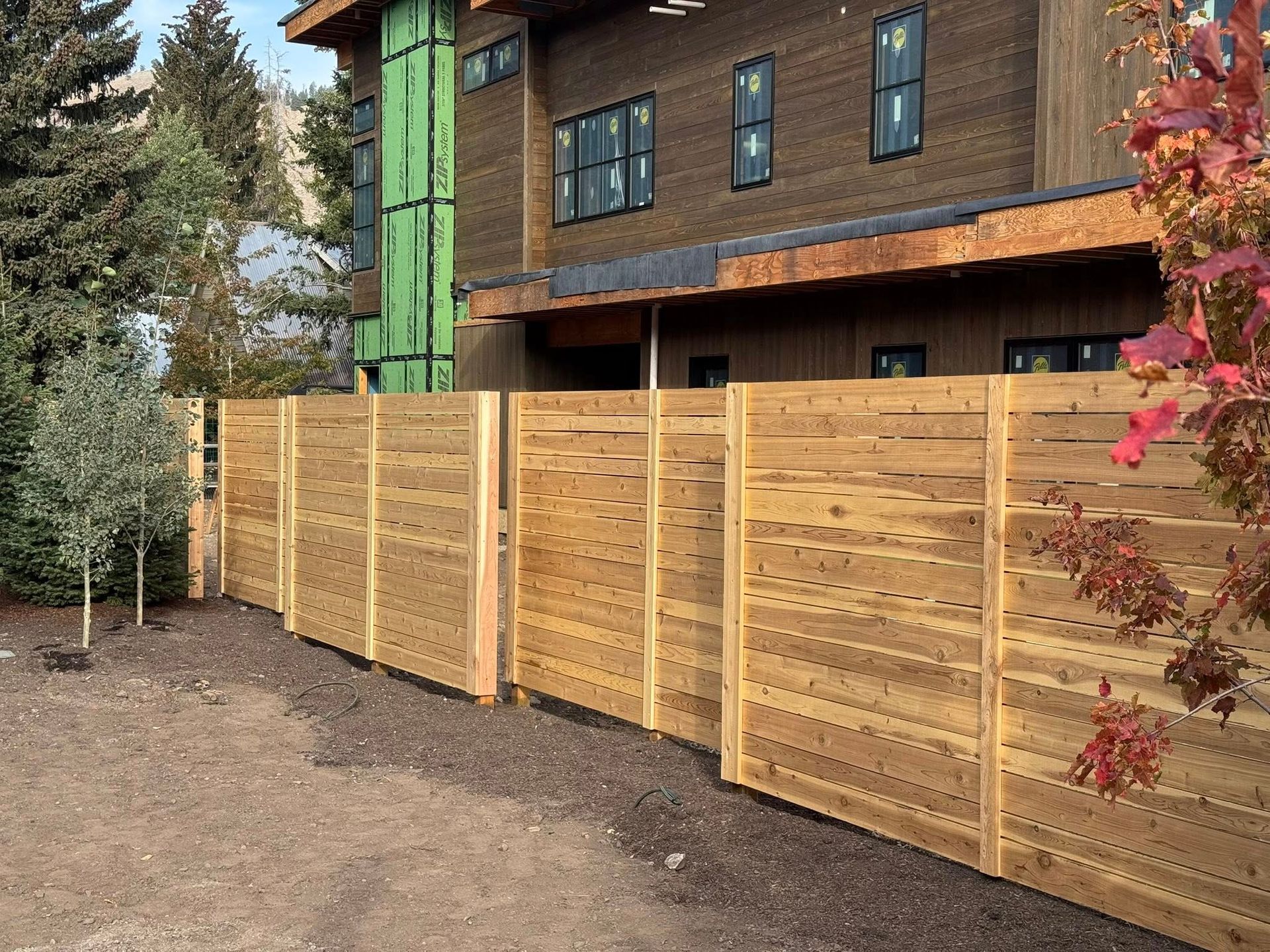 Wooden fence in front of a modern house, dirt ground, trees, and blue sky in background.