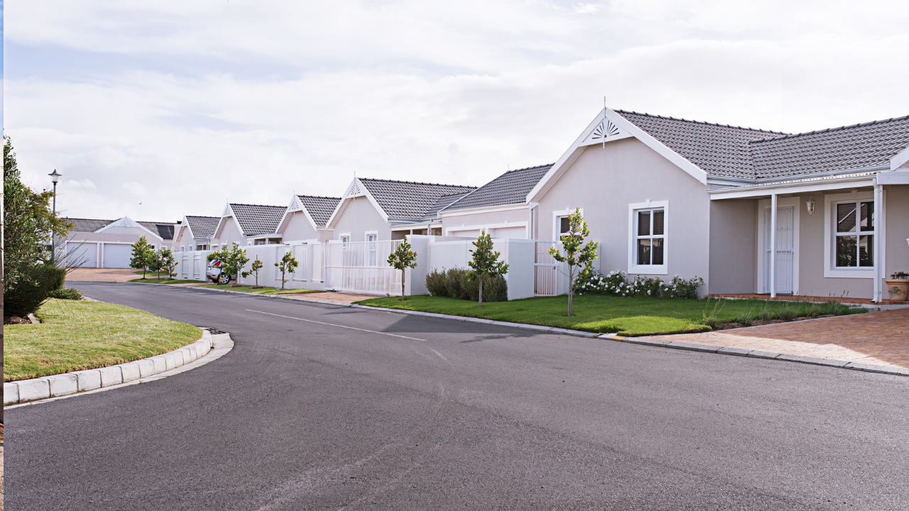 Row of light-colored houses with white fences and grey roofs along a curving asphalt road under a cloudy sky.