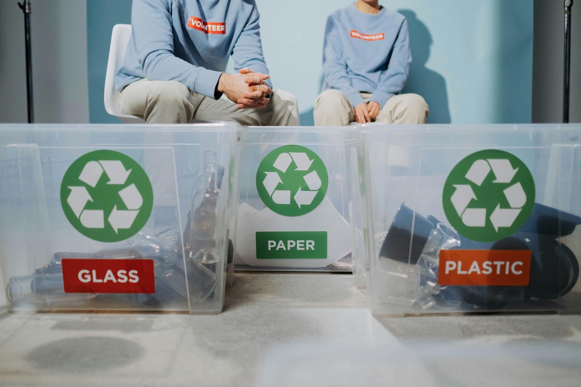 People sitting behind recycling bins labeled for glass, paper, and plastic.