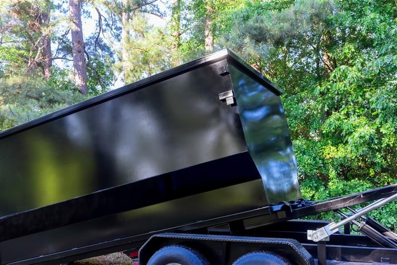 Black dump trailer, tilted, with a wooded background.