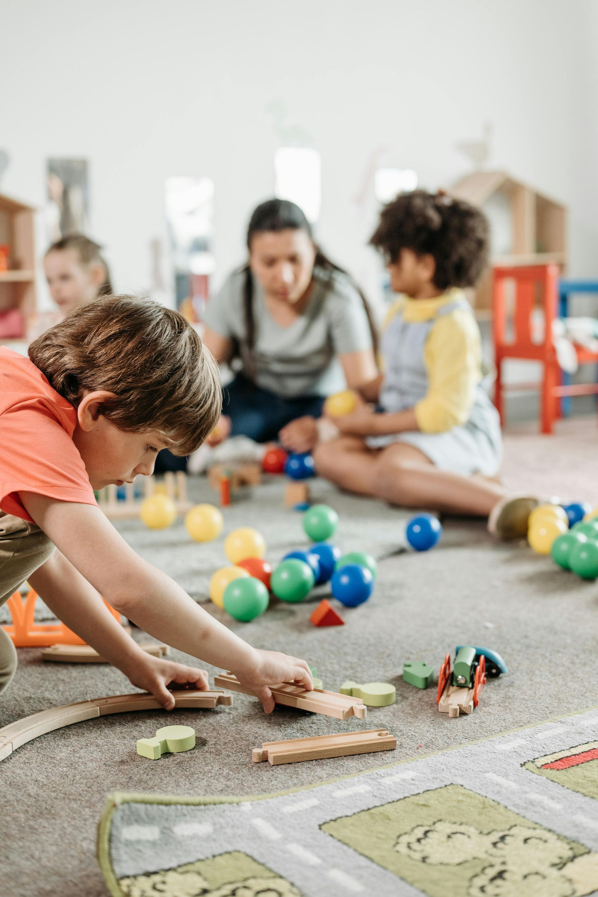 Dans une pièce lumineuse, une institutrice et trois enfants jouent avec des balles colorées, des blocs et des rails de train miniatures sur un sol recouvert de moquette.