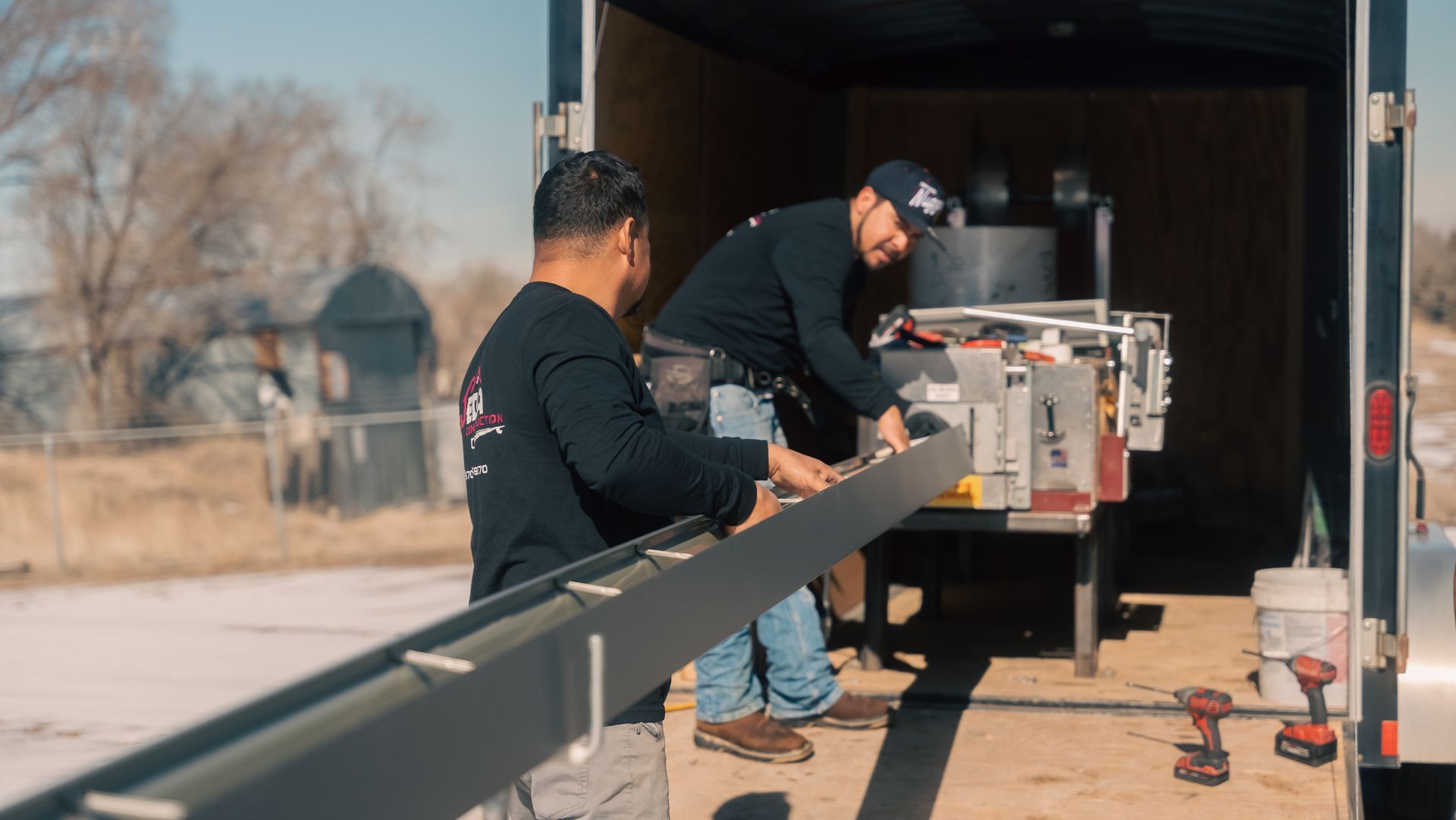 Two men are working on a piece of metal in front of a trailer.