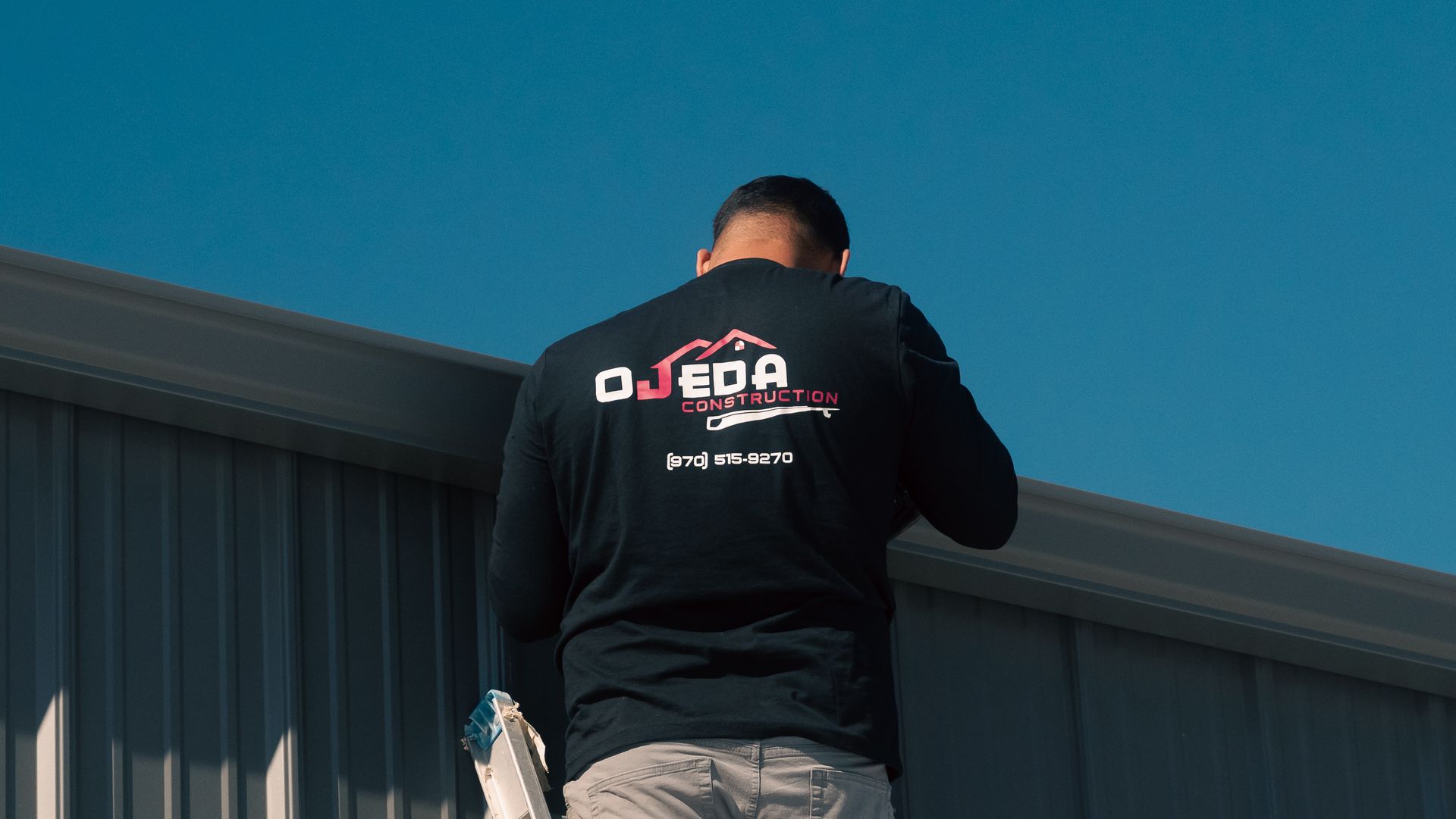 A man wearing a black shirt with the word ojeda on the back is standing in front of a building.