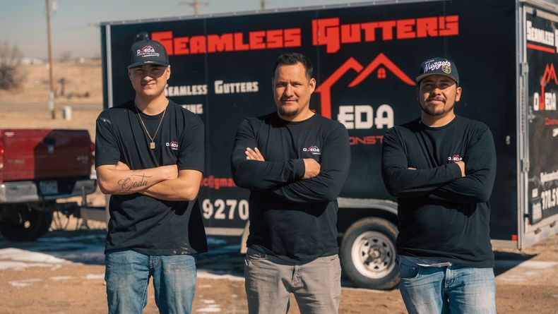 Three men are standing in front of a trailer that says seamless gutters.