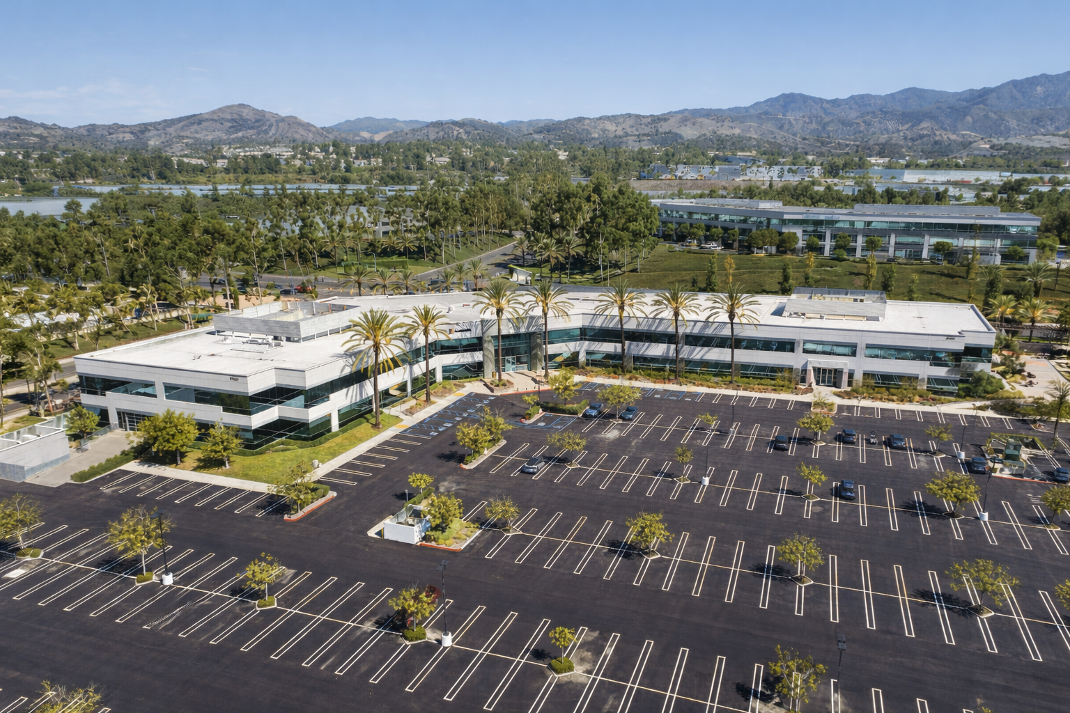 Aerial view of a low-rise office building with a large parking lot, surrounded by greenery and distant mountains.