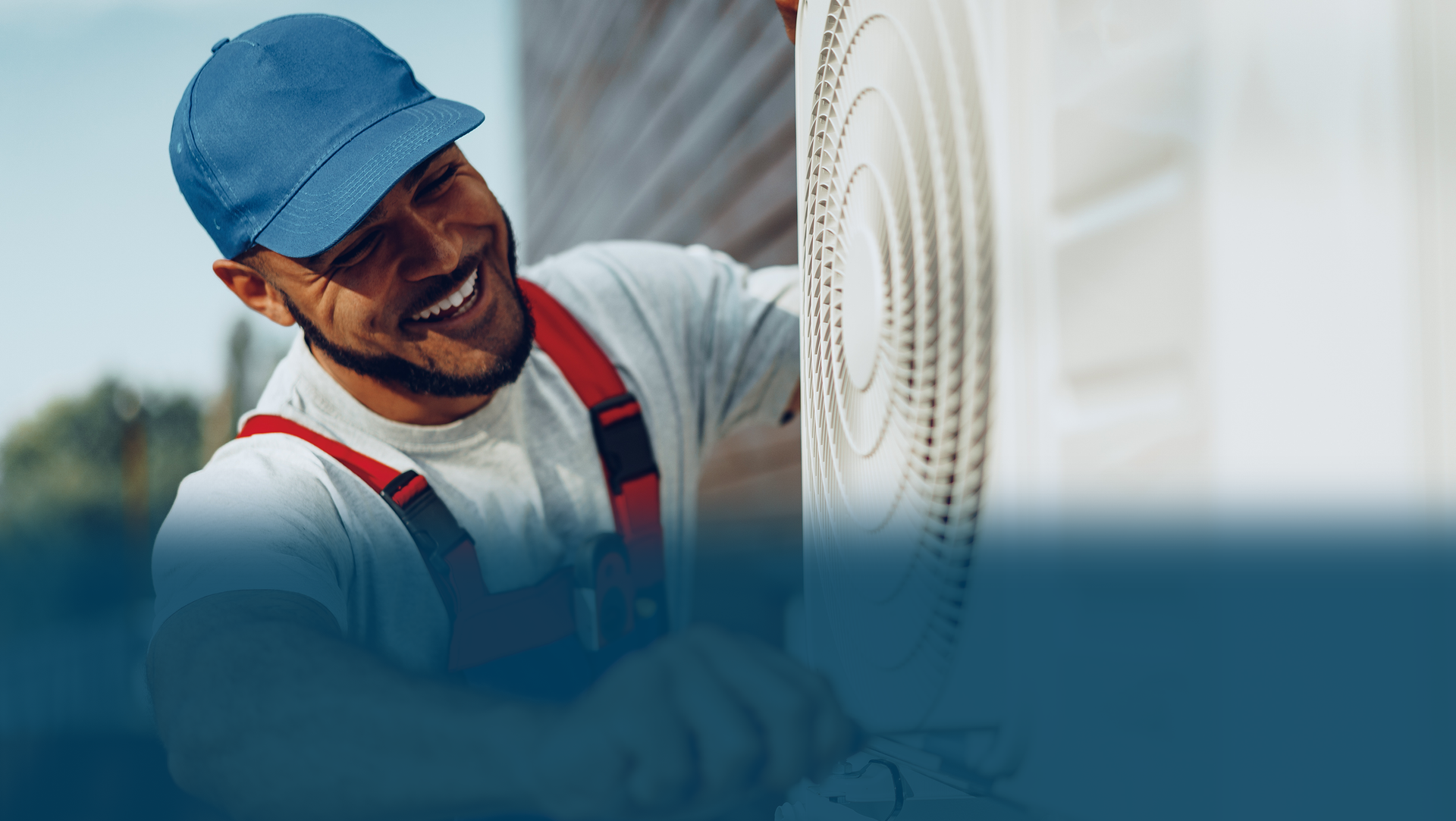 HVAC technician smiling while working on an outdoor AC unit; wearing a blue cap and overalls.
