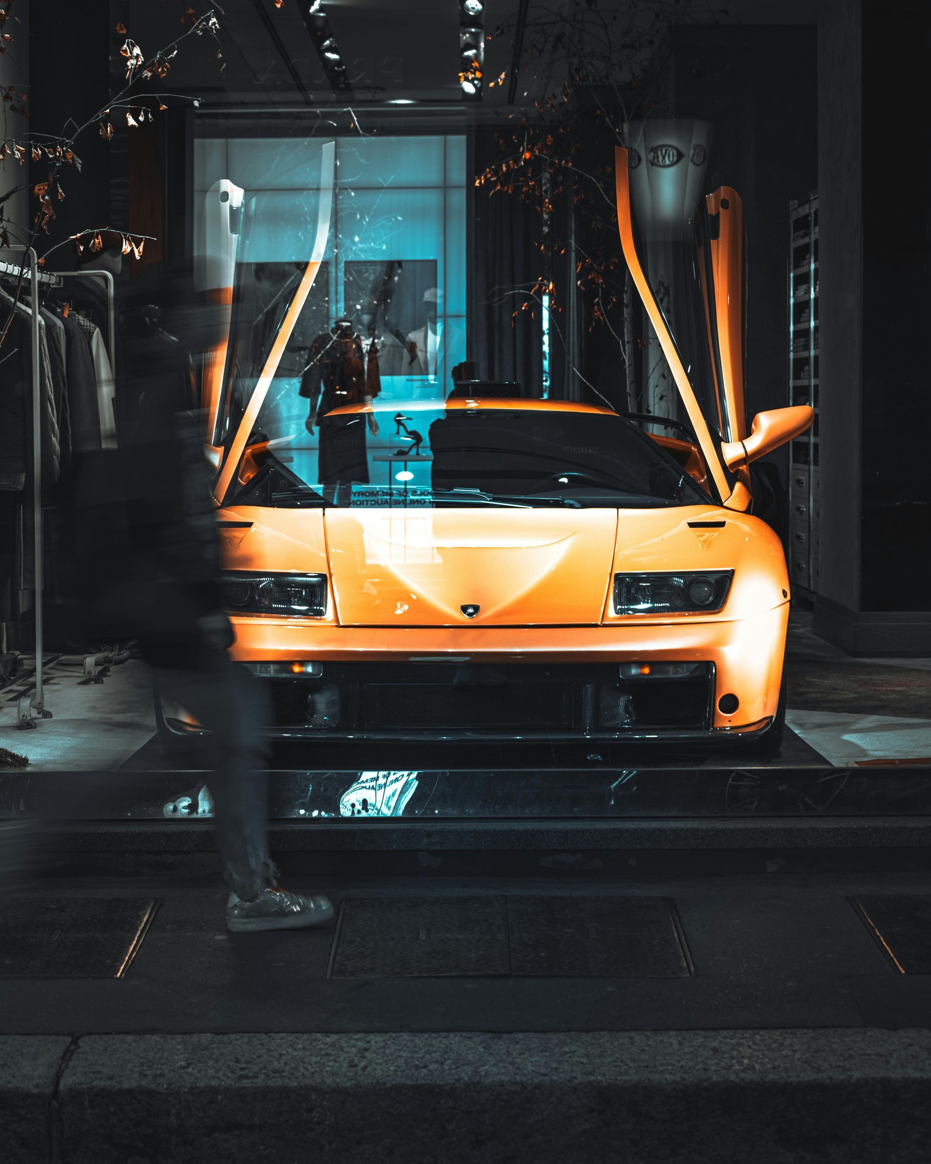 Orange Lamborghini with open doors, parked outside a store. Blurred person walks by.