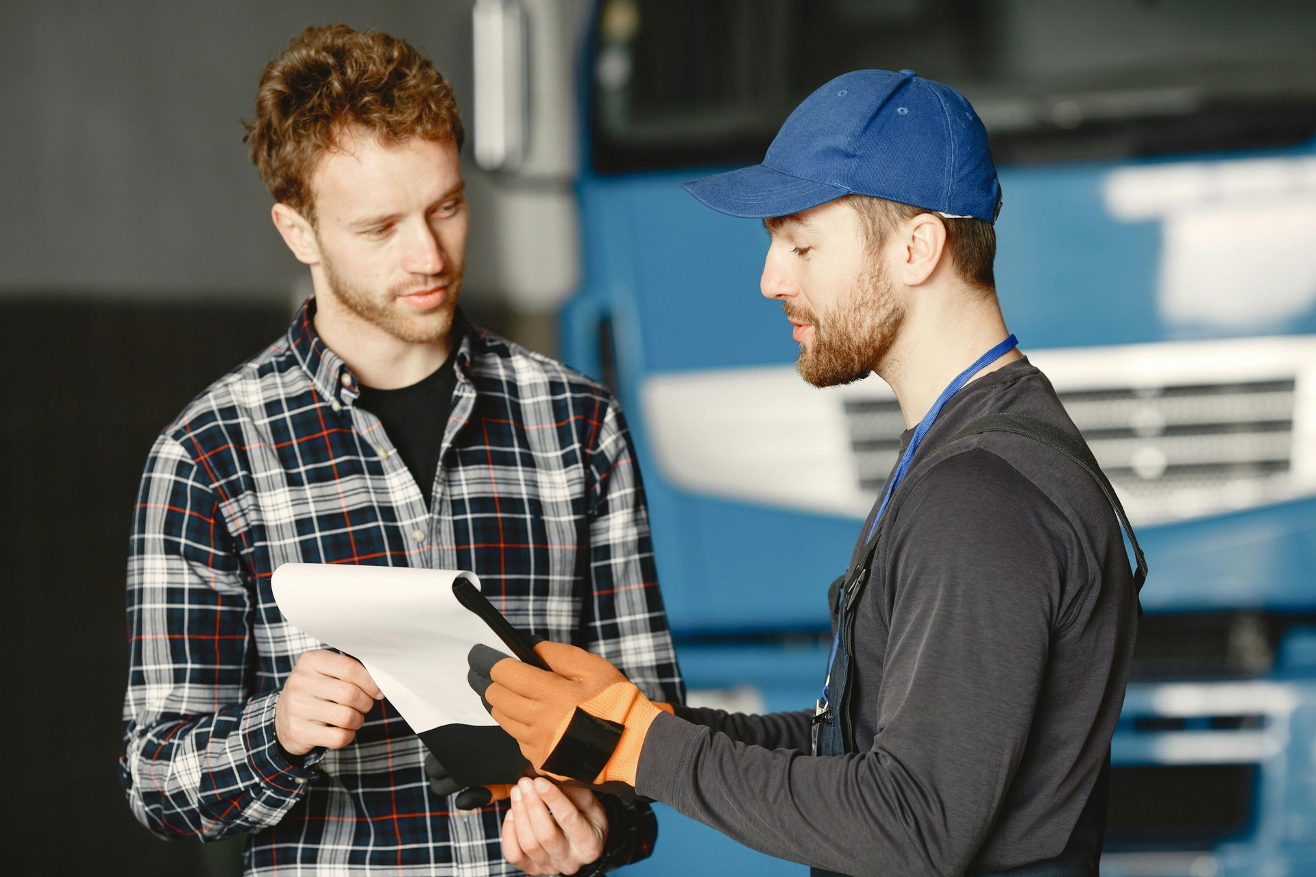 Man in plaid shirt reviews paperwork with a worker in blue cap and work gloves near a blue truck.