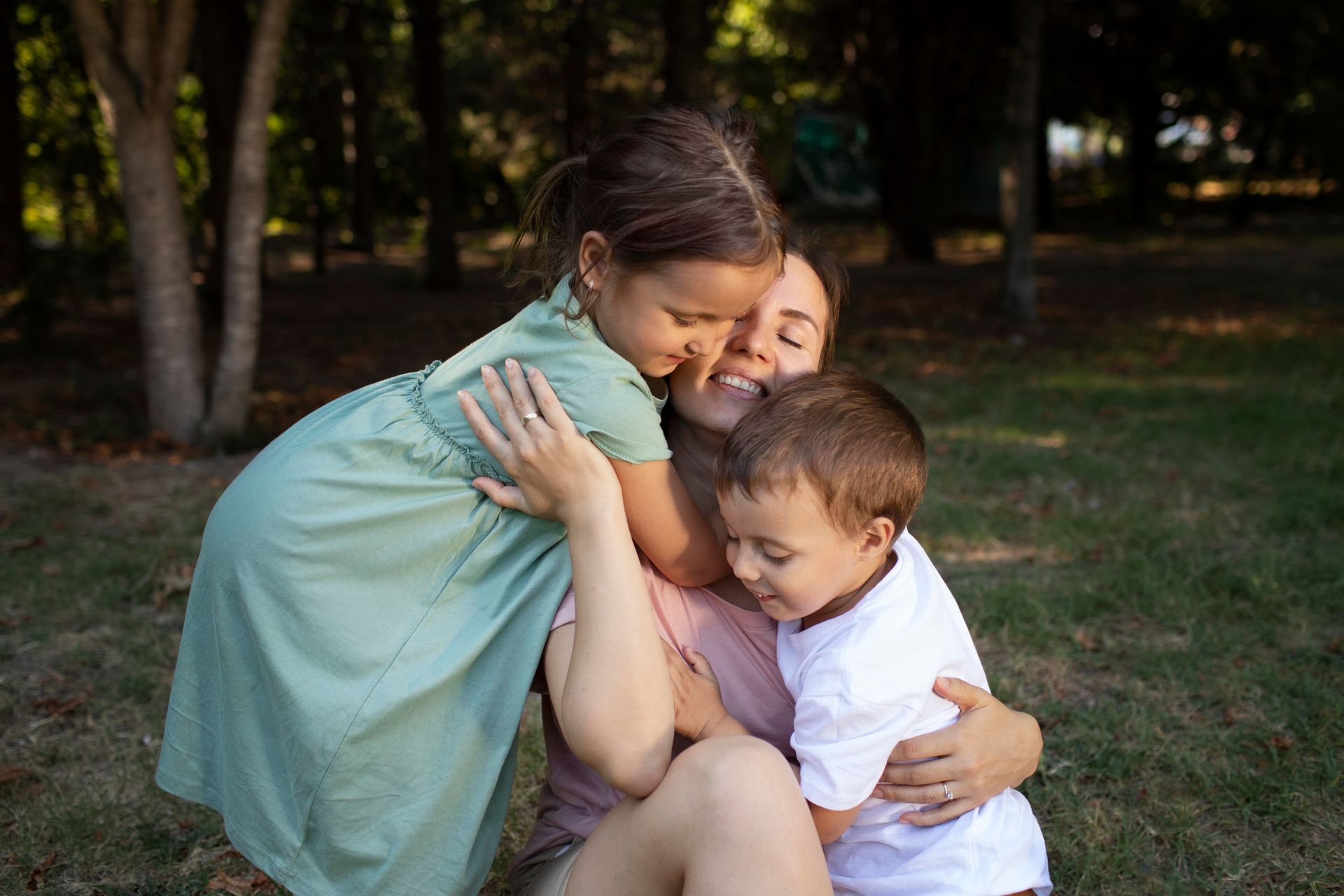 Woman smiles while embracing two young children in a sunny park.
