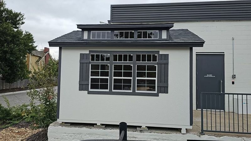 A white shed with a black roof and shutters is sitting in front of a building.