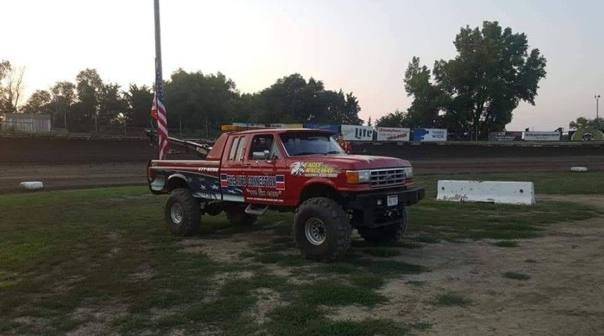 Red lifted pickup truck with an American flag on the field of a racetrack.