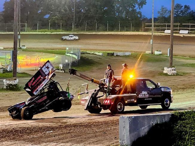 A sprint car being towed by a tow truck at a dirt race track at dusk.