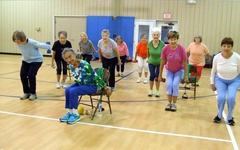 Group of people exercising in a gymnasium. Some standing, others using chairs. All are bending and smiling.