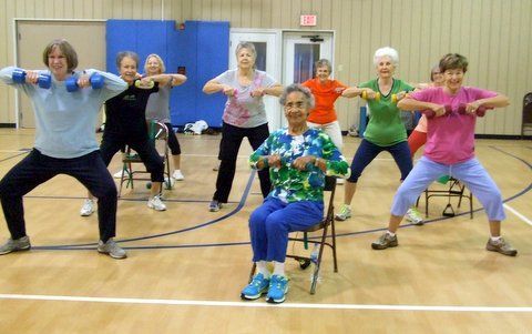 Group of people exercising with weights in a gym. Some standing, some seated.