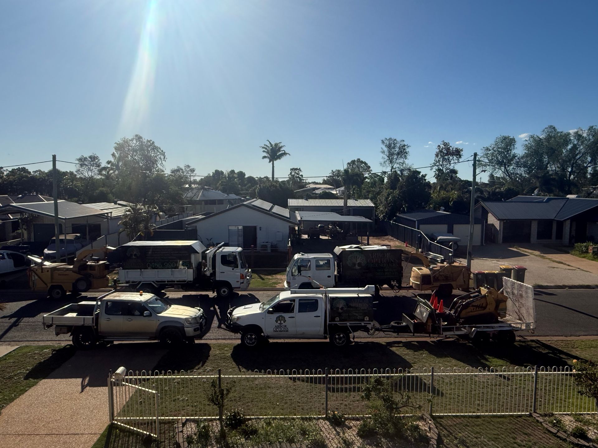 A row of service trucks and a small excavator on a trailer parked on a residential street under a sunny sky.