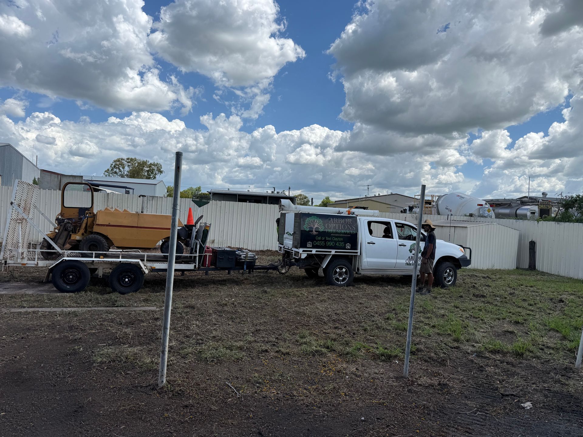 A white pickup truck towing a flatbed trailer with yellow construction equipment on a dirt lot under a cloudy sky.