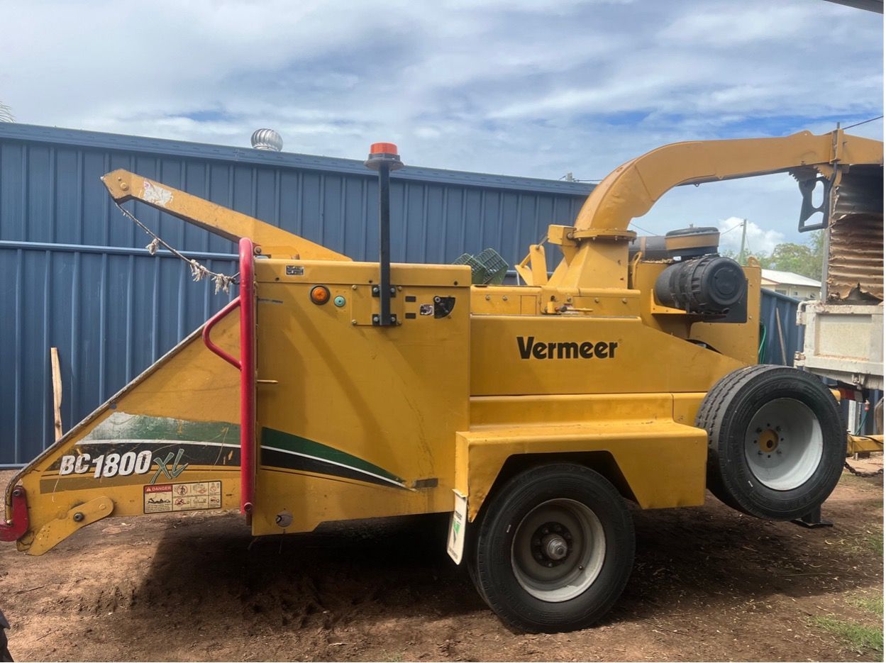 A yellow Vermeer BC1800 wood chipper parked on gravel in front of a blue metal wall.
