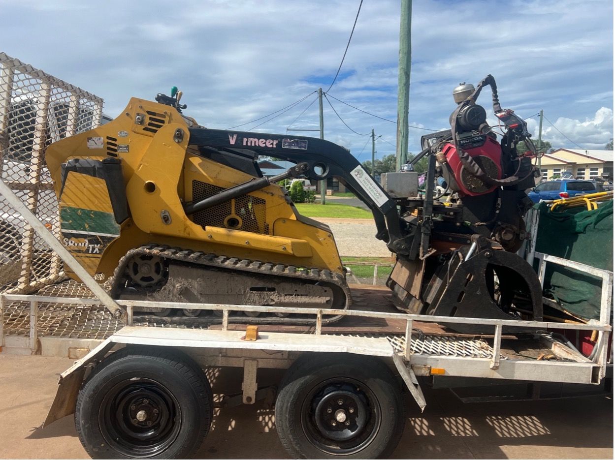 A yellow compact track loader with a tree-mulching attachment is loaded on a flatbed trailer outdoors.