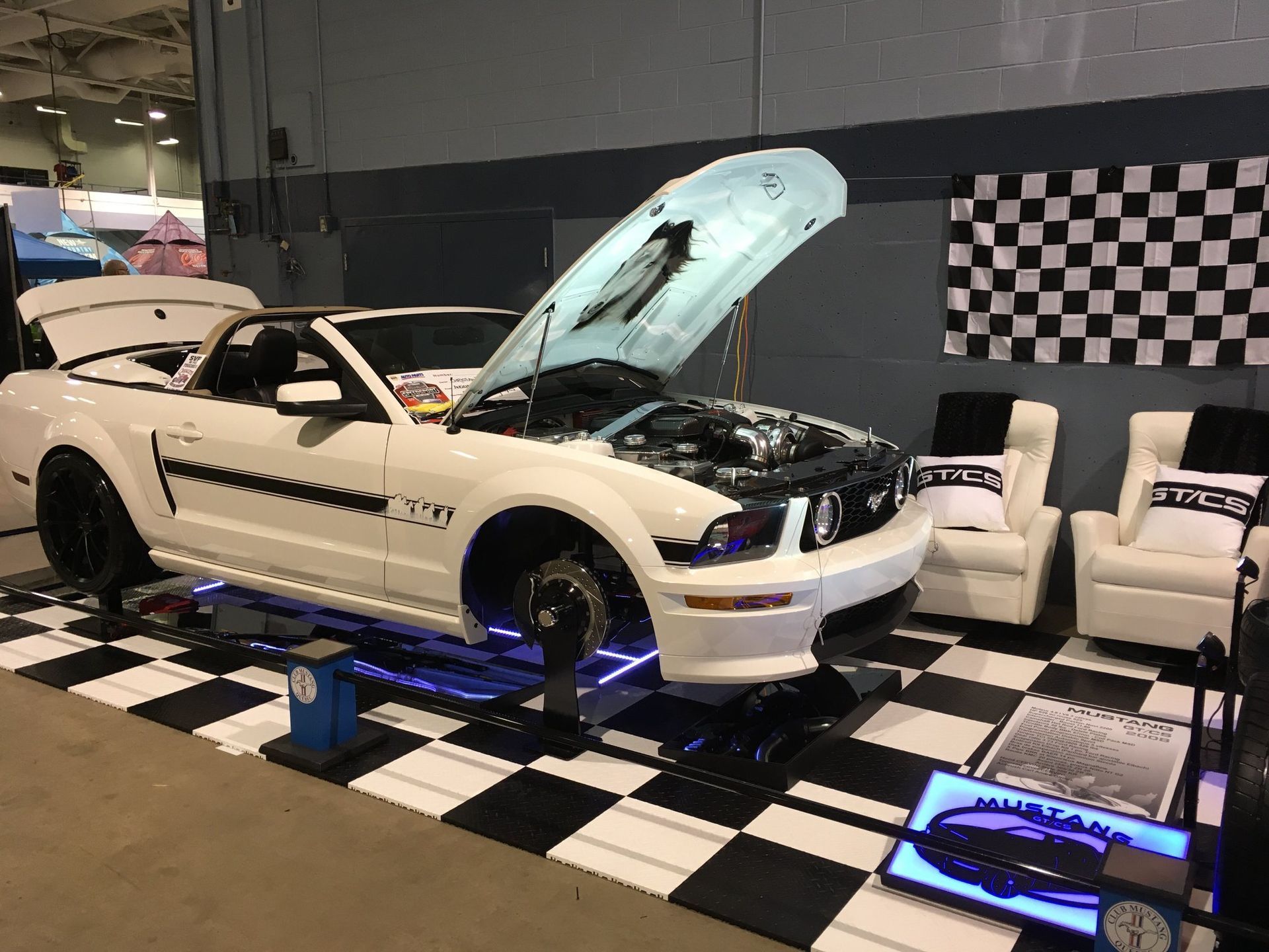 Une Ford Mustang décapotable blanche exposée lors d'un salon automobile, capot ouvert, intérieur visible, feux bleus en dessous.