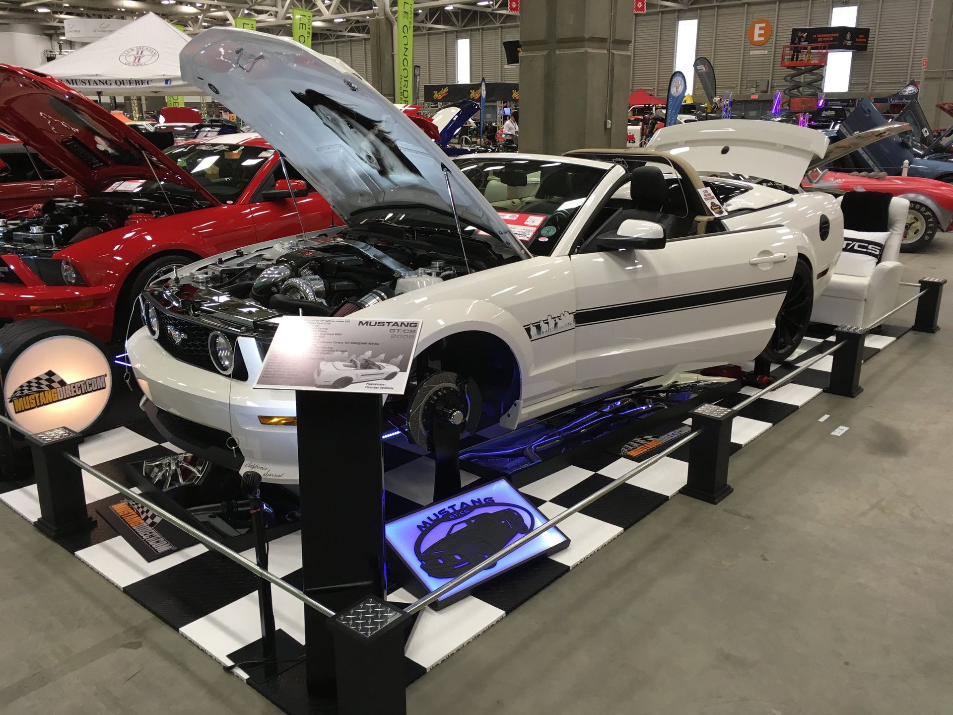 Ford Mustang décapotable blanche exposée lors d'un salon automobile, capot et coffre ouverts, plancher à damier.