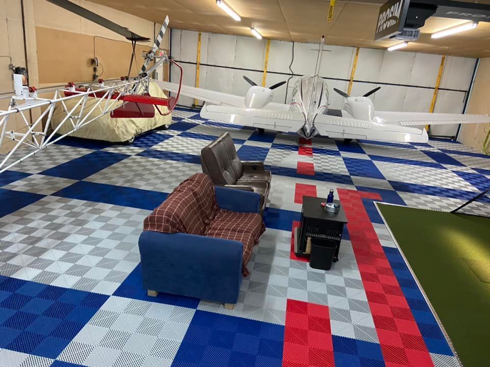 Hangar à avions avec un avion bimoteur, un canapé et une zone recouverte de moquette rouge sur un sol carrelé.