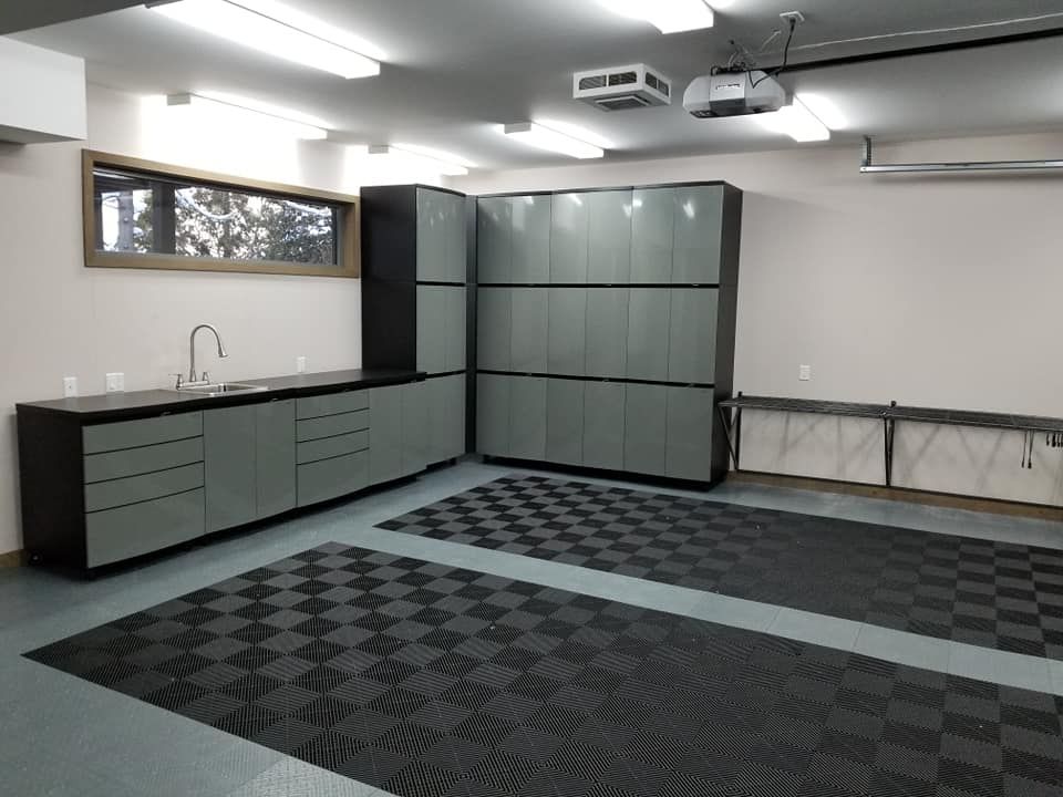 Garage avec placards gris, évier et sol à damier. Murs blancs, éclairage fluorescent.
