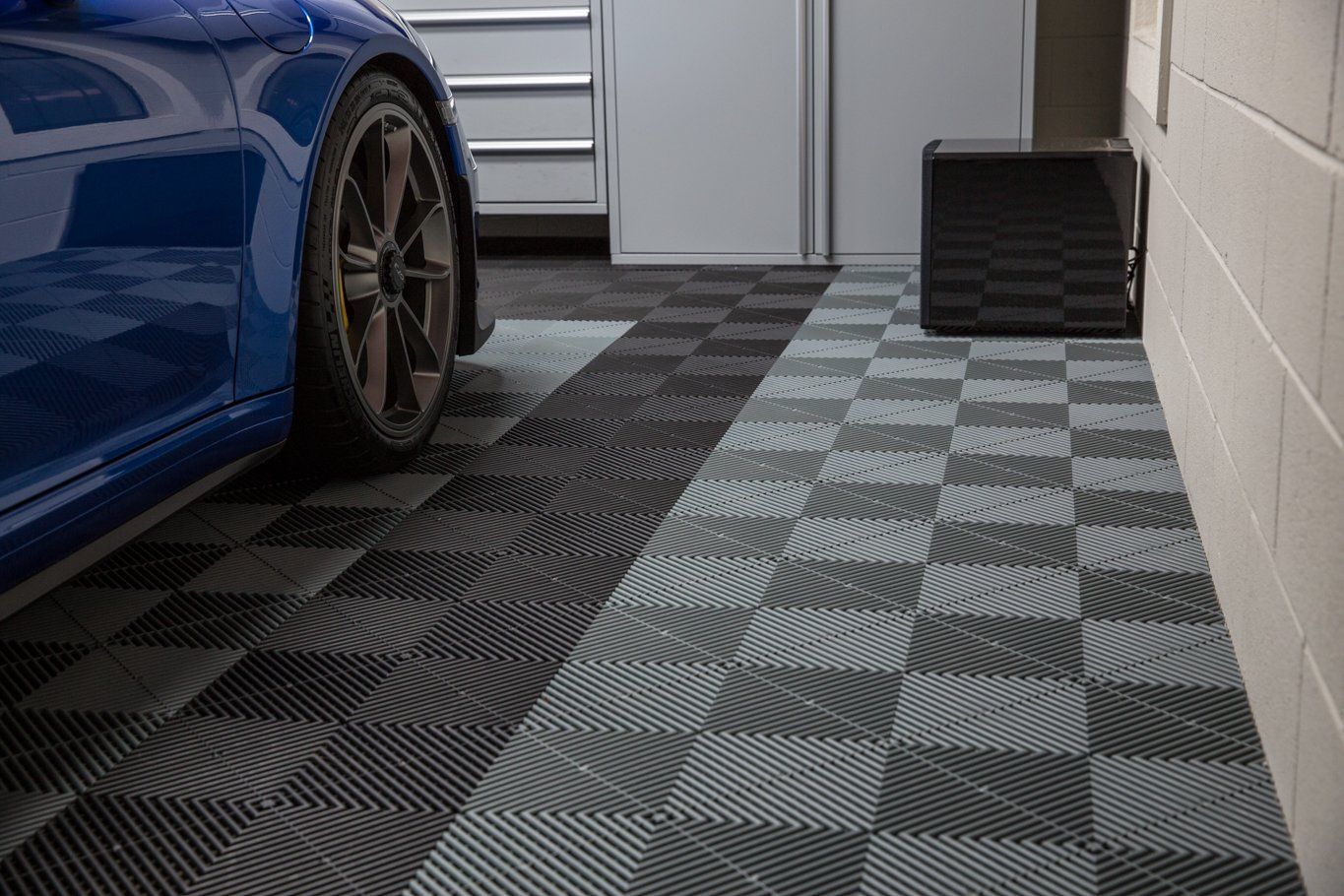 Une voiture bleue est garée dans un garage au sol carrelé de carreaux à damier et à rainures. Une enceinte noire est adossée à un mur blanc.