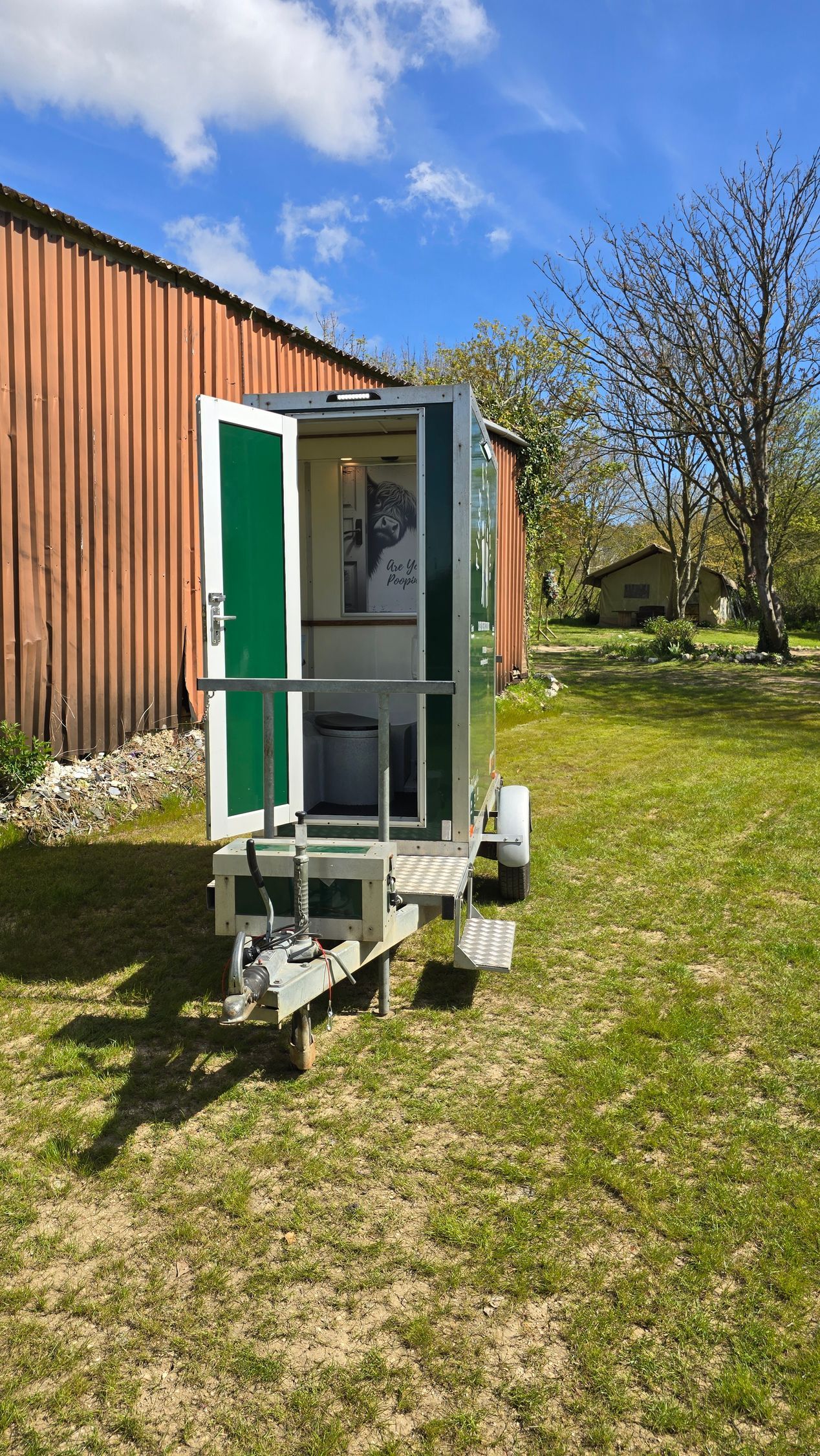 Small white utility trailer with a green door parked on grass beside a red barn under a blue sky.