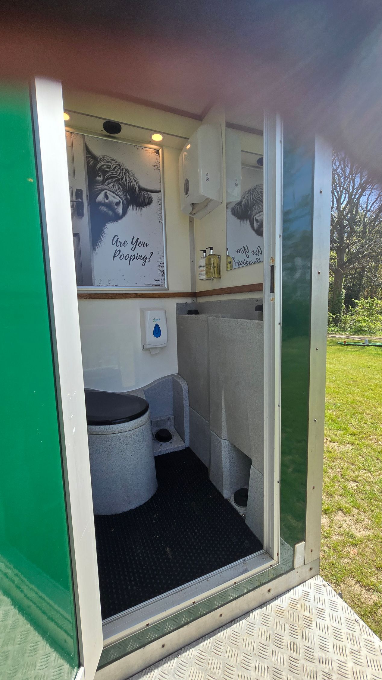 Open portable toilet with white sink, black floor mat, and green door, set outdoors near grass.
