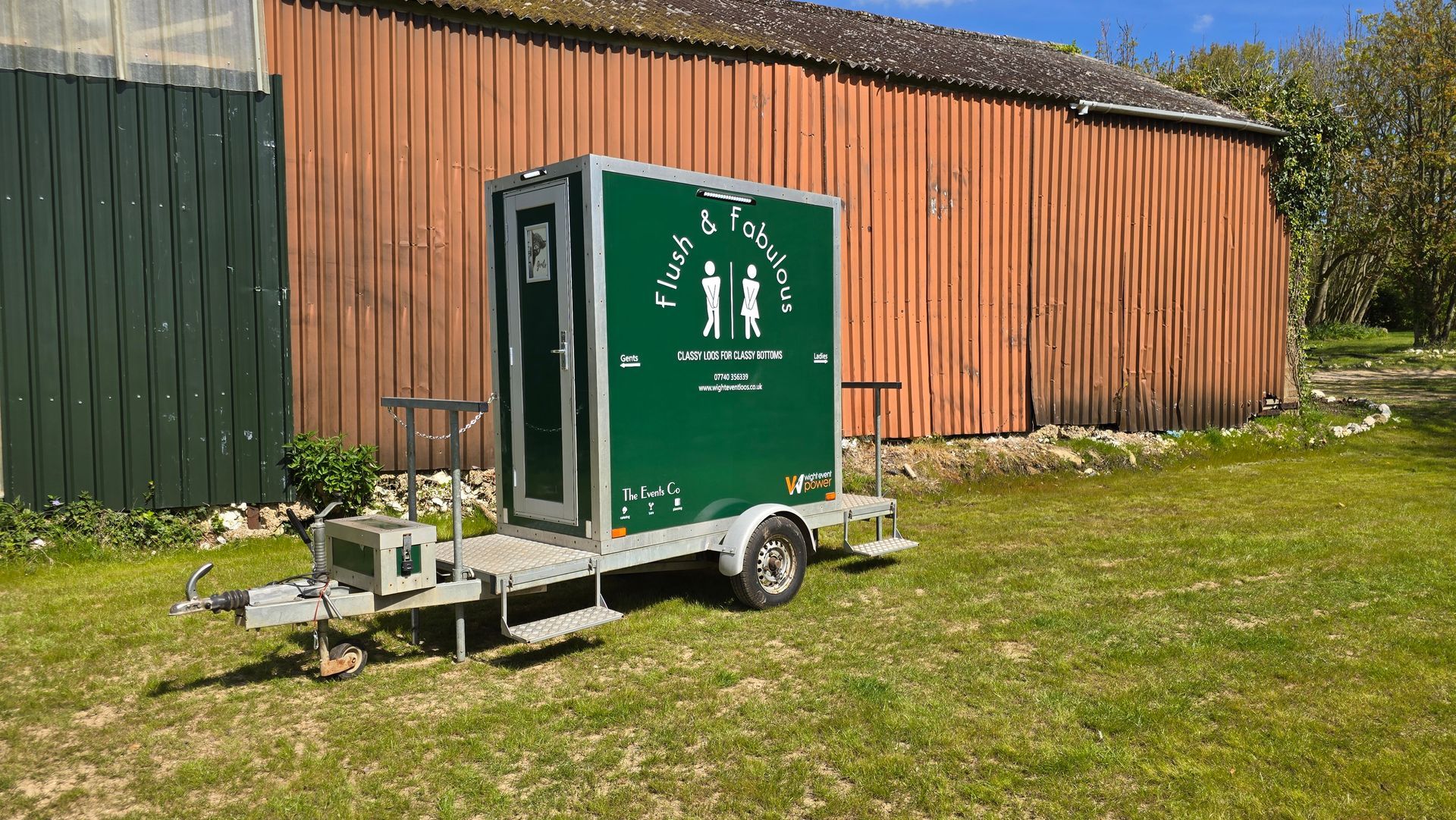 Green portable toilet trailer parked on grass beside a wooden fence and building.