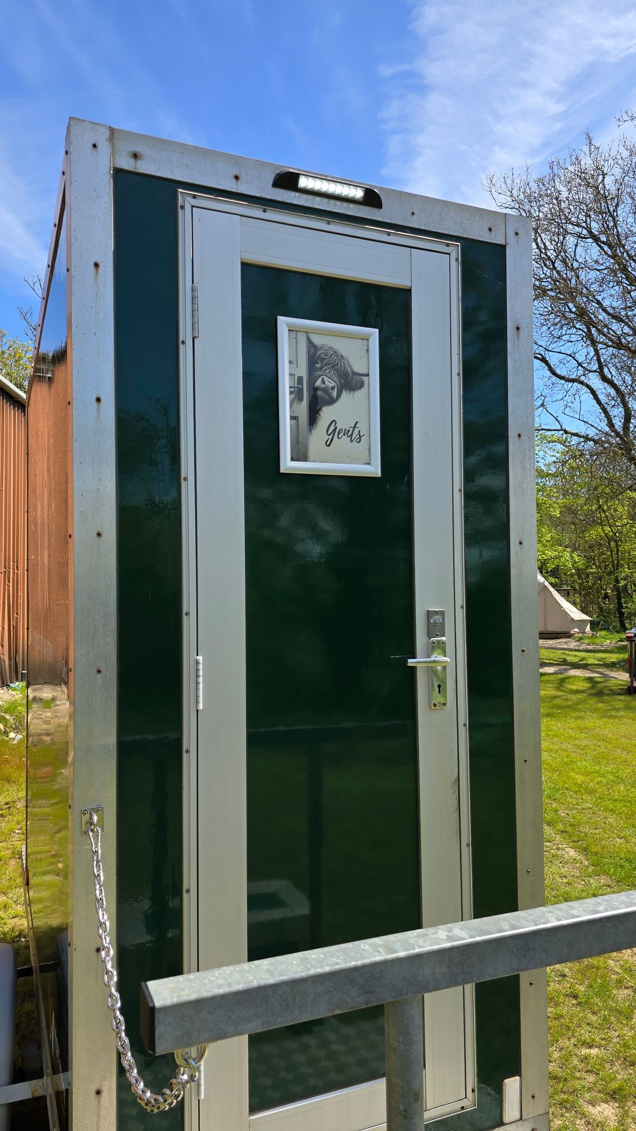 Green portable toilet with silver trim and a small window, outdoors beside a fence.