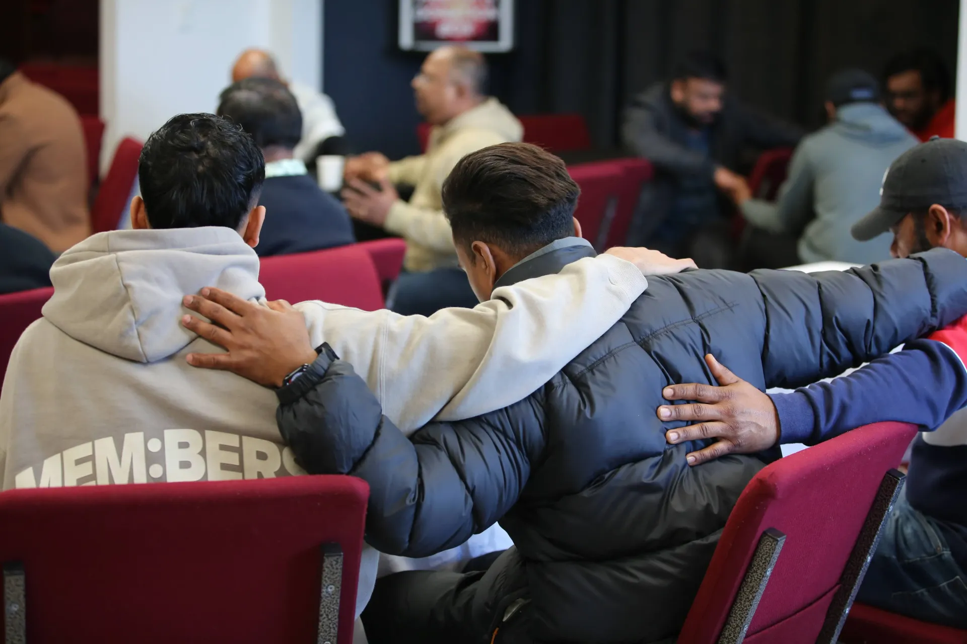 Two men with arms around each other's shoulders, sitting in a room with other people.