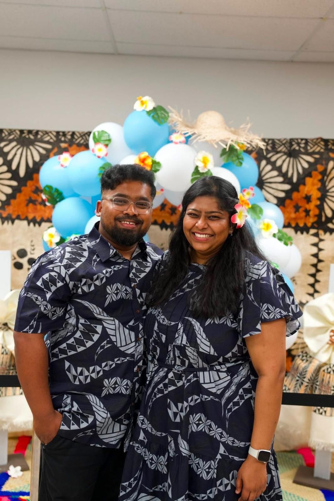 Couple in matching blue patterned shirts smile, posing in front of balloons and decorations.