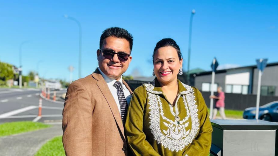 Man in a suit and woman in green smiling near a road with buildings and a clear blue sky.