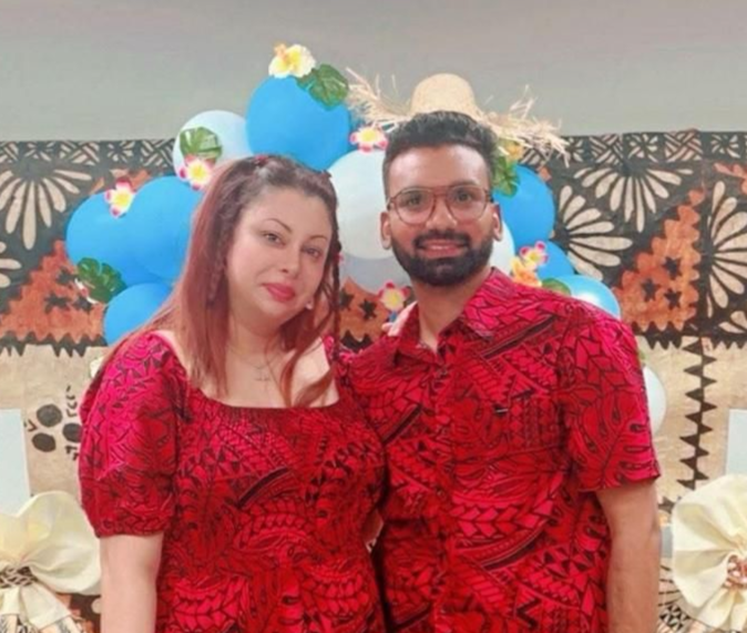Couple in matching red patterned shirts, smiling, posing in front of blue balloons and a patterned backdrop.