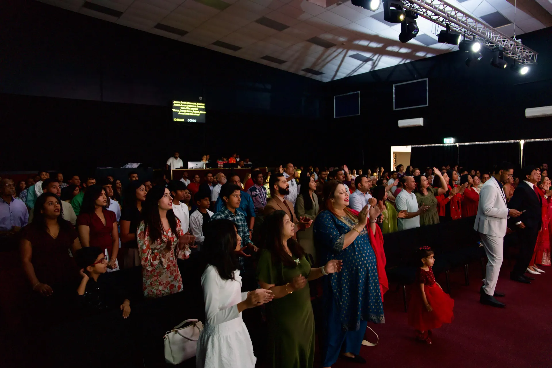 Large crowd in an auditorium, many with hands raised, led by a person in a white suit on a stage with lights.