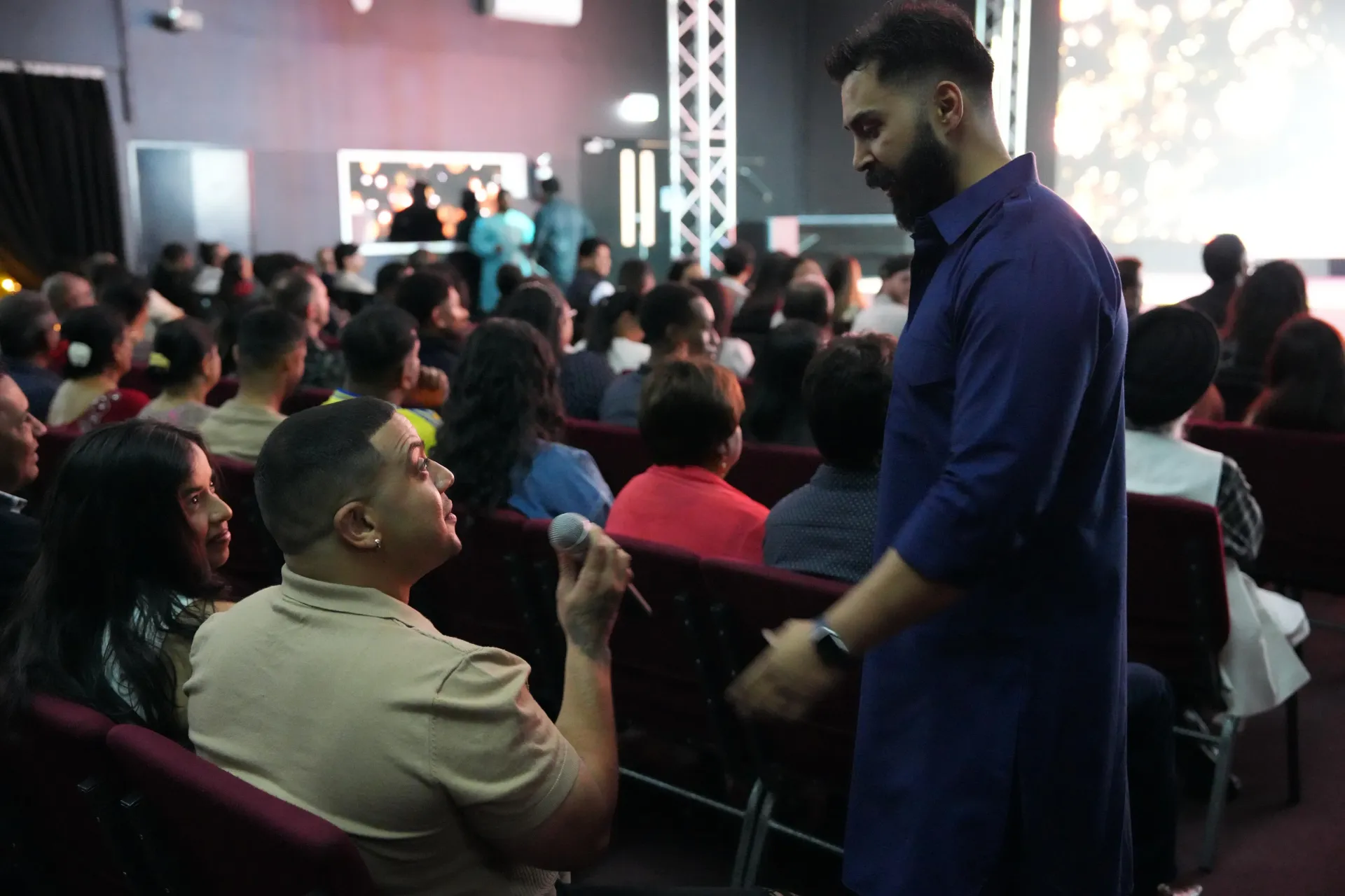 Man in blue shirt speaks with another man holding a microphone in a church setting with audience.