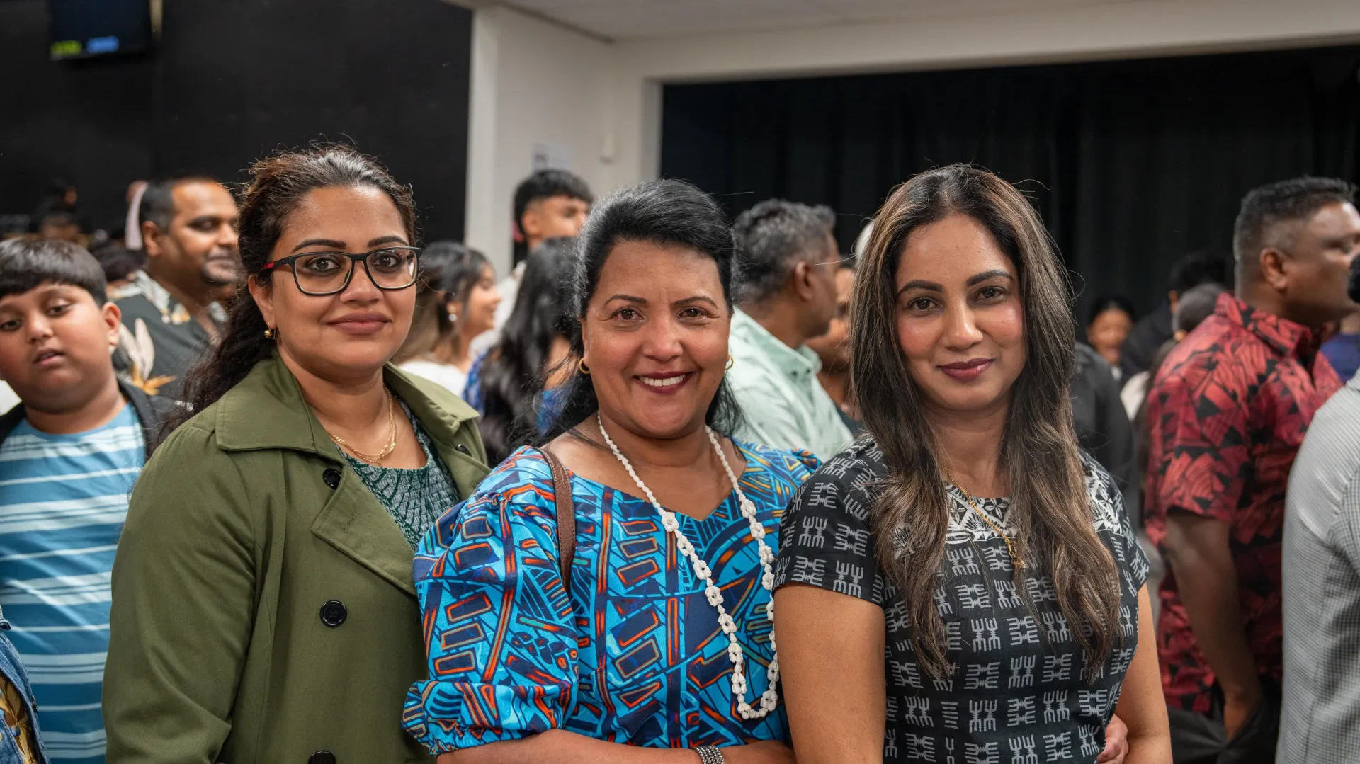 Three women smiling at an event; other people in the background.