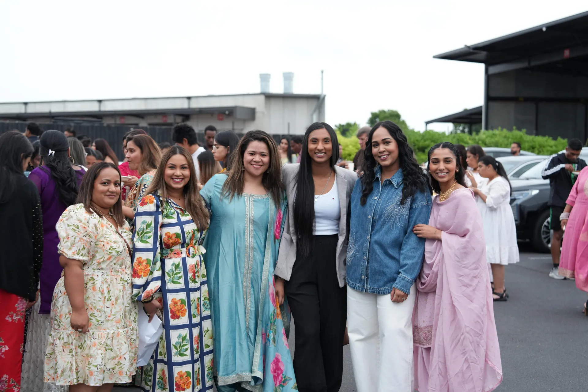 Group of people in colorful outfits posing outdoors. Buildings and parked cars in background.