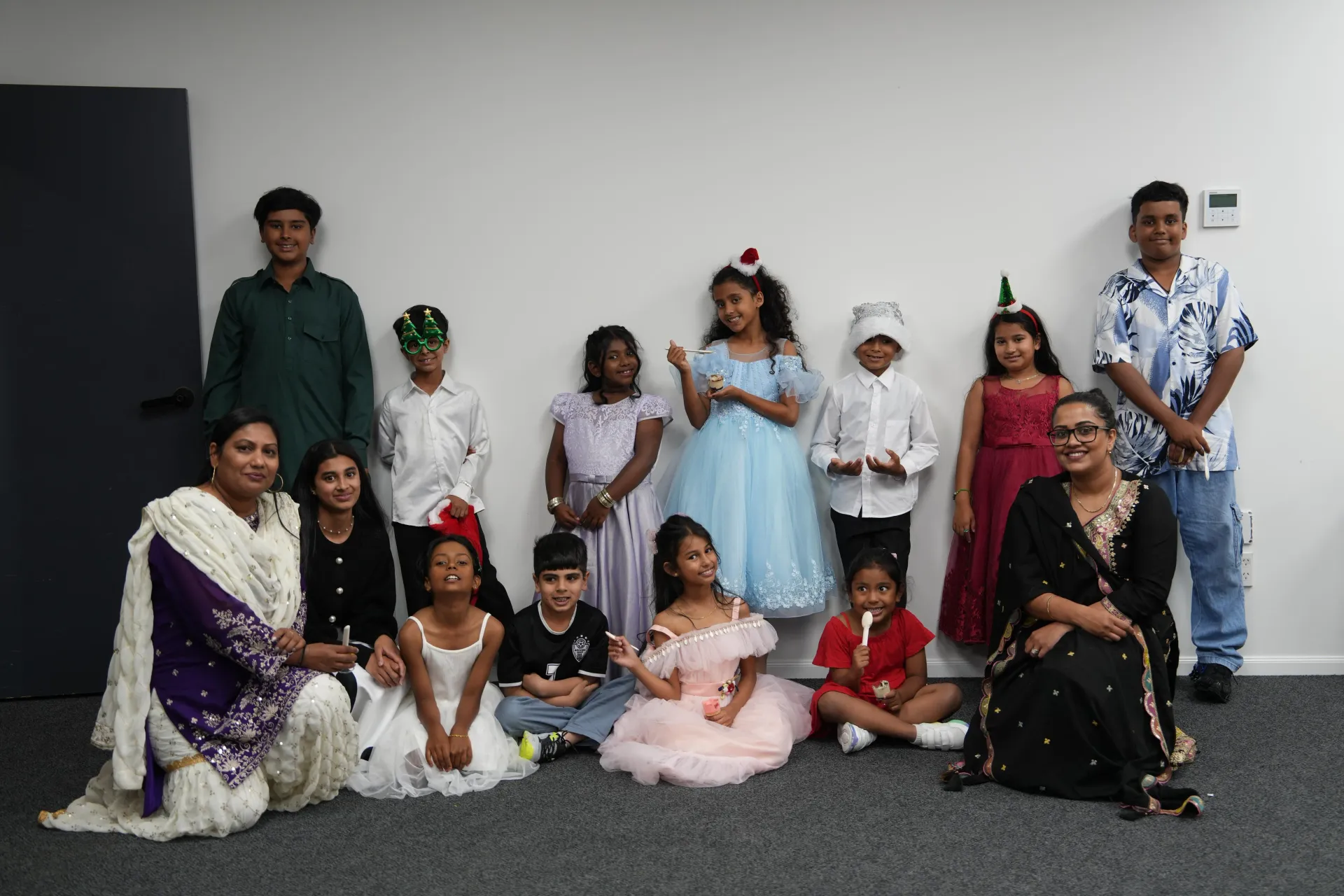 Group of people, including children, pose for a photo in a room with a white wall. Some wear festive headwear.