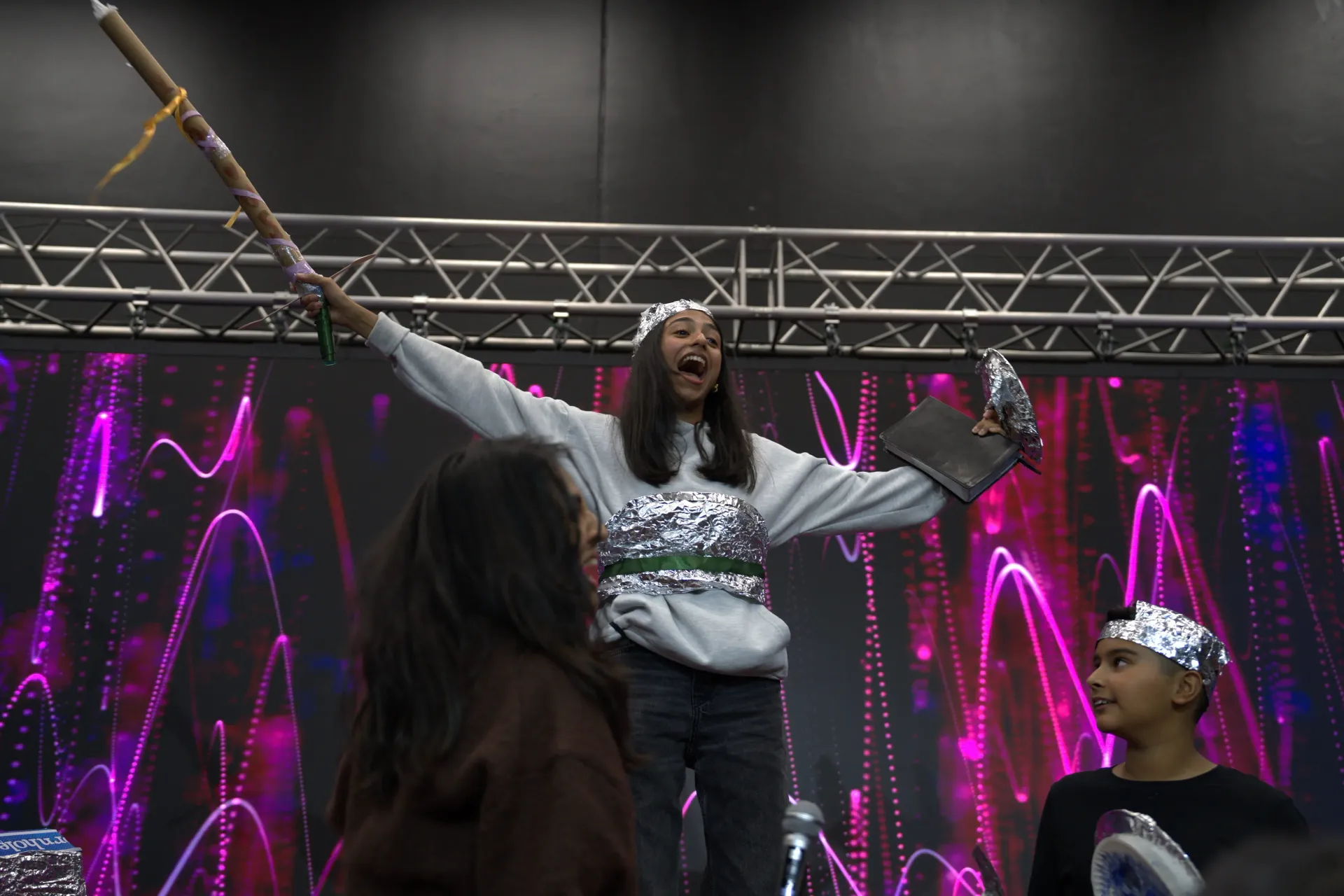 Person on stage with arms raised, holding stick. People in the audience, one with foil crown. Bright pink and purple lights in the background.