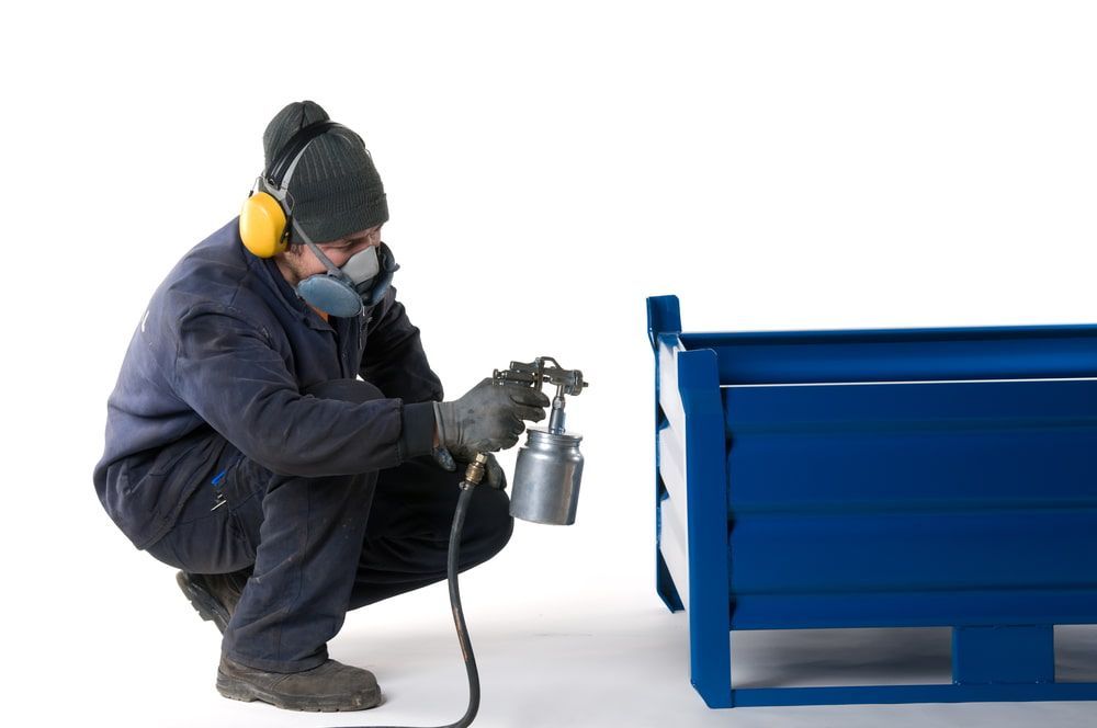 A Man Wearing a Mask and Ear Muffs is Spray Painting a Blue Box — NHM Painting & Projects Pty Ltd in Ciccone, NT