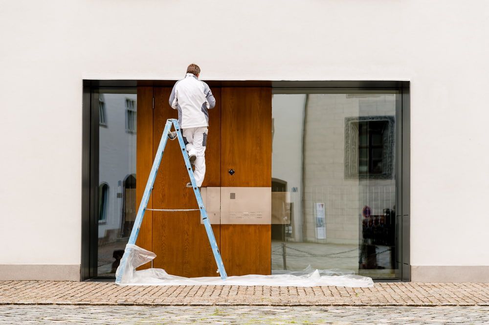 A Man is Standing on a Ladder Painting a Door — NHM Painting & Projects Pty Ltd in Ciccone, NT