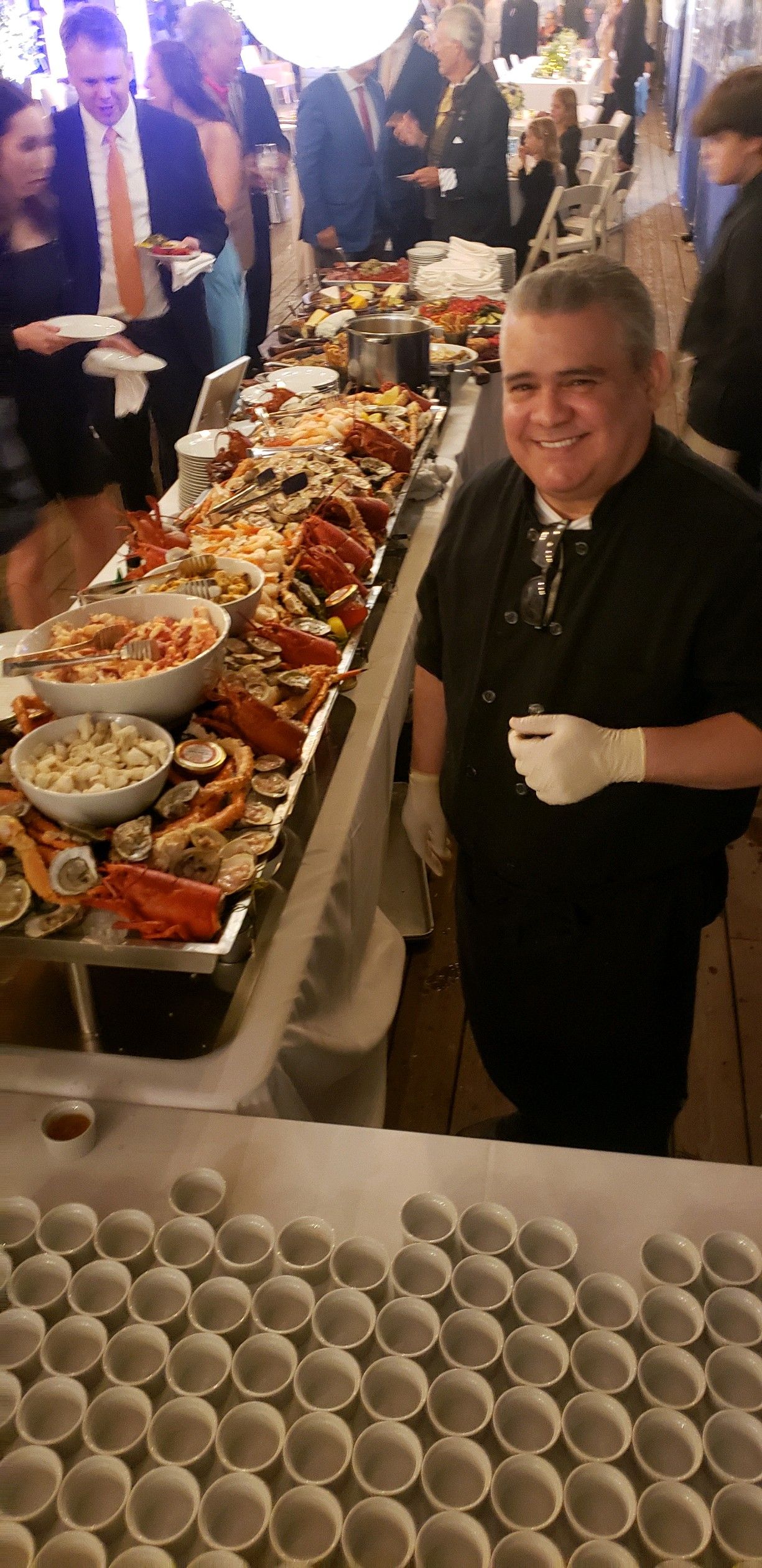 A chef smiles, standing beside a long seafood buffet table. Guests in formal attire are in the background.