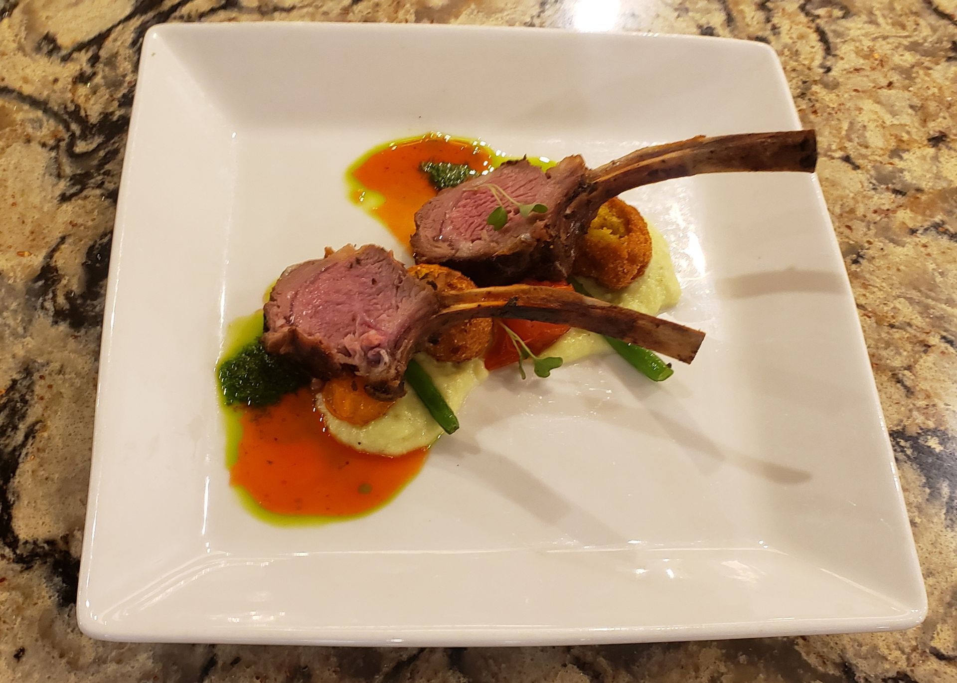 Rack of lamb with sauce and vegetables on a square white plate, set on a patterned countertop.