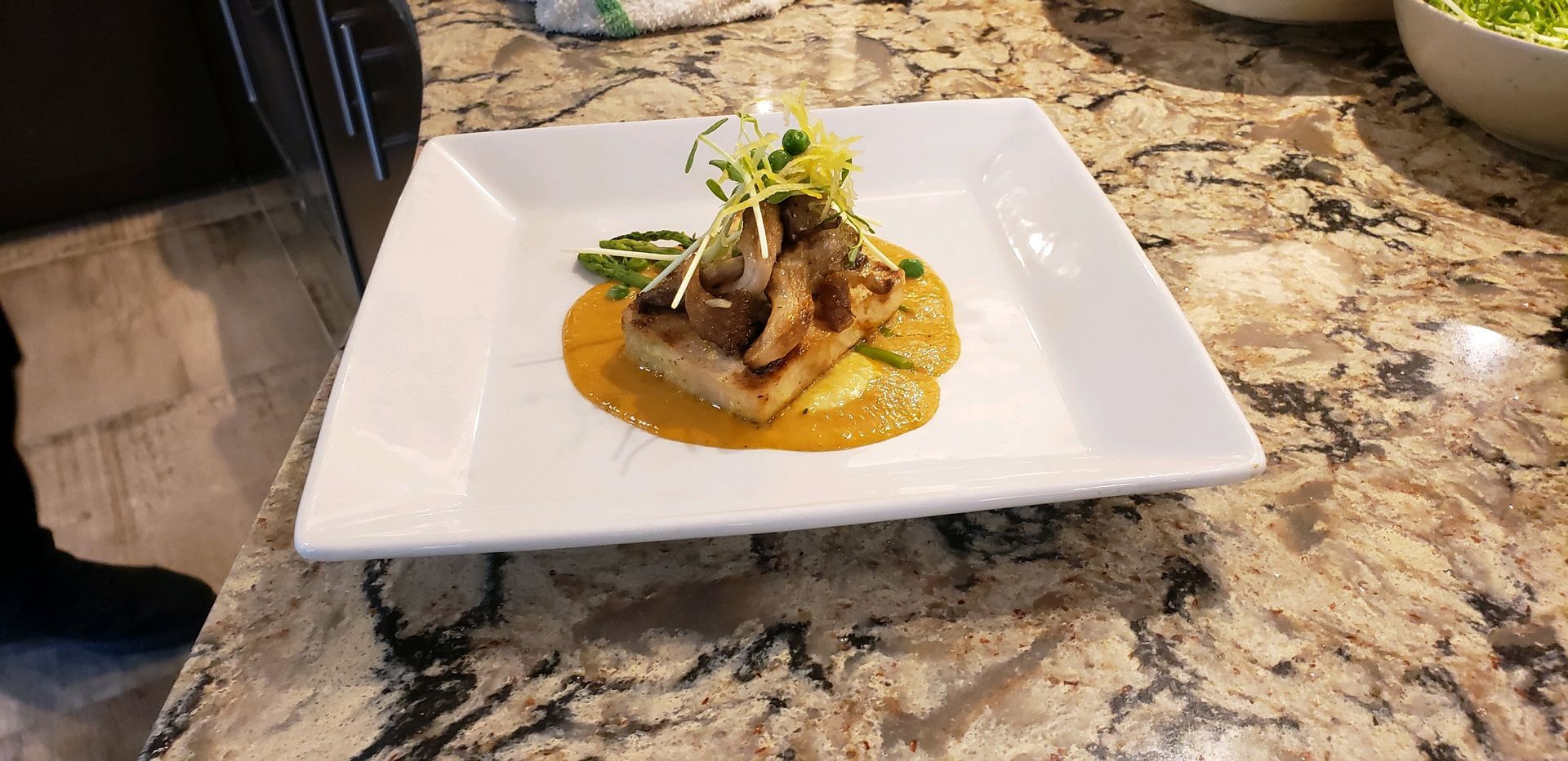 Square white plate with gourmet dish of fish, sauce, and vegetables.  It is on a granite countertop.